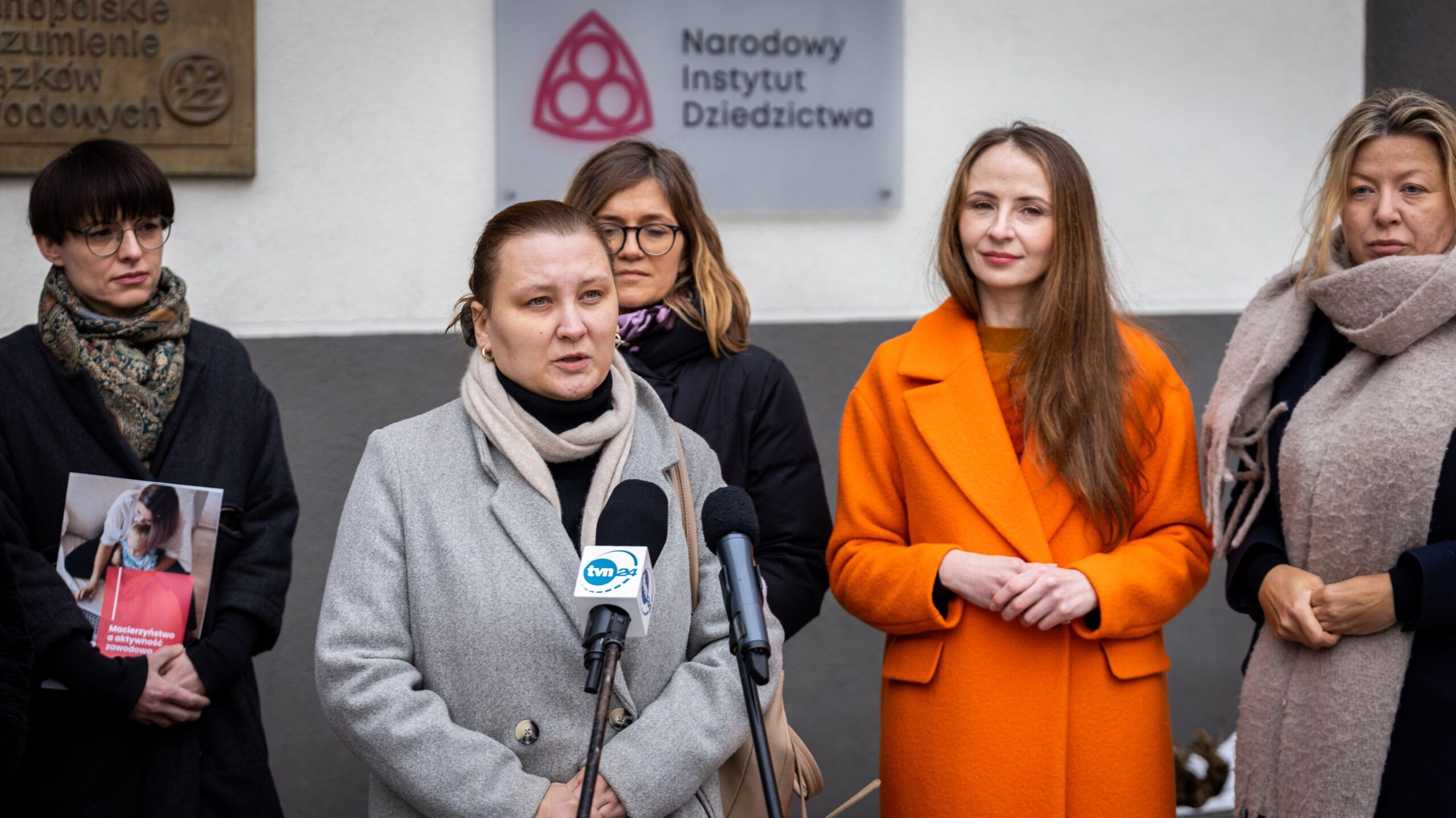 Na zdjęciu widoczne są cztery kobiety stojące przed siedzibą Narodowego Instytutu Dziedzictwa, podczas wydarzenia, na którym jedna z nich przemawia do mikrofonu.