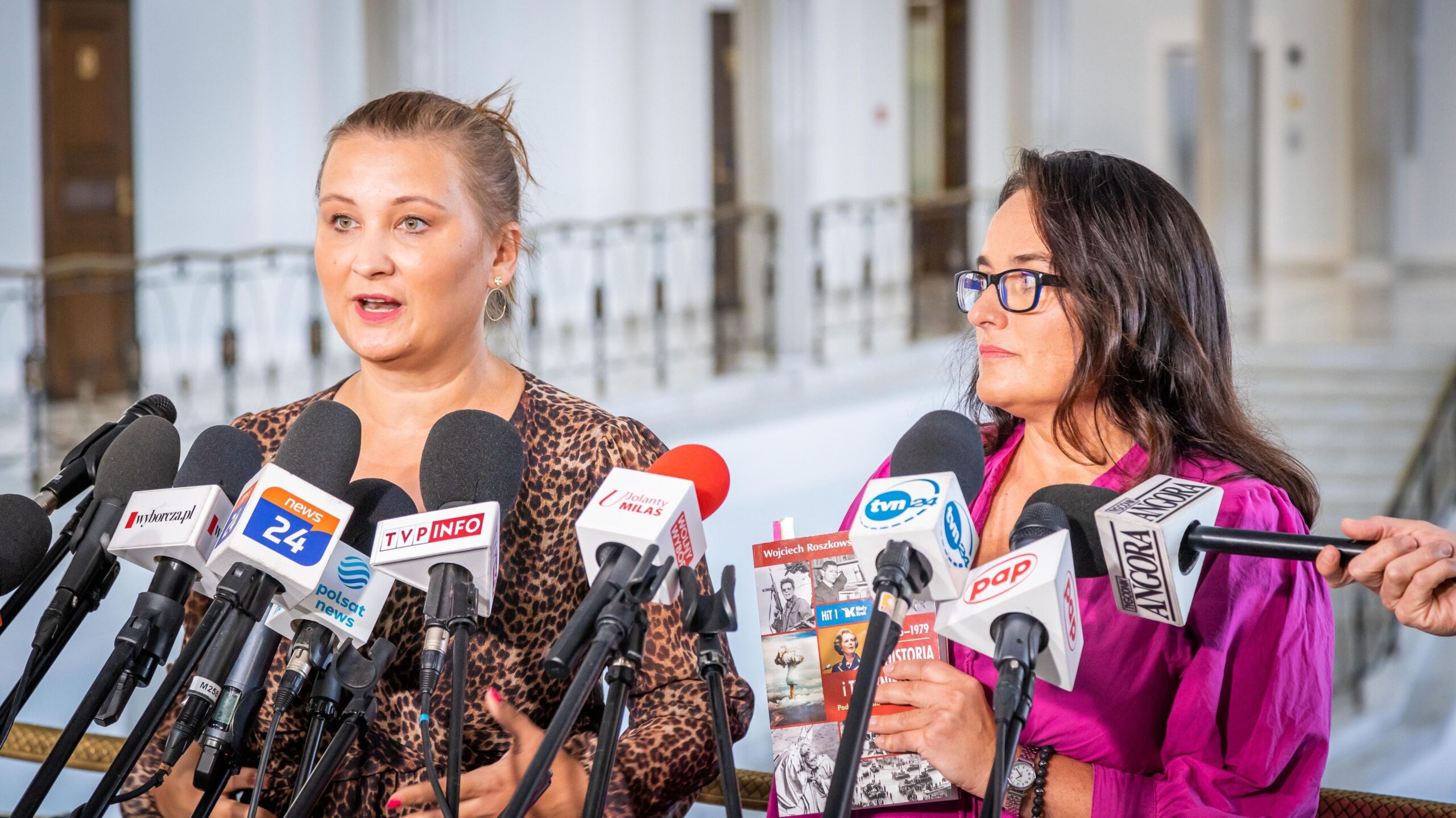 Na zdjęciu dwie kobiety stoją przy mównicy otoczonej mikrofonami i rozmawiają w trakcie konferencji prasowej.