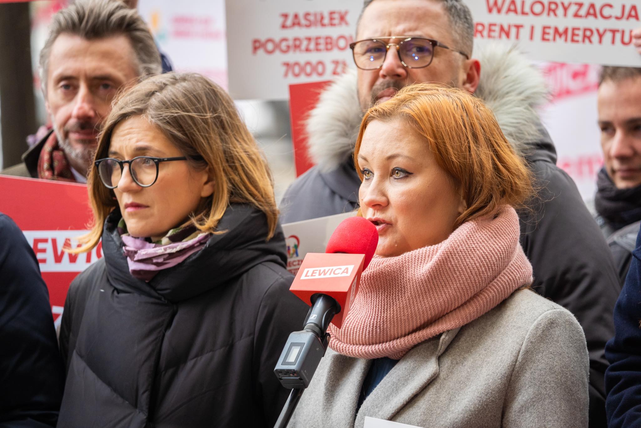 Na zdjęciu widać kobietę z mikrofonem, która przemawia podczas demonstracji, obok niej stoją inni uczestnicy trzymający transparenty.