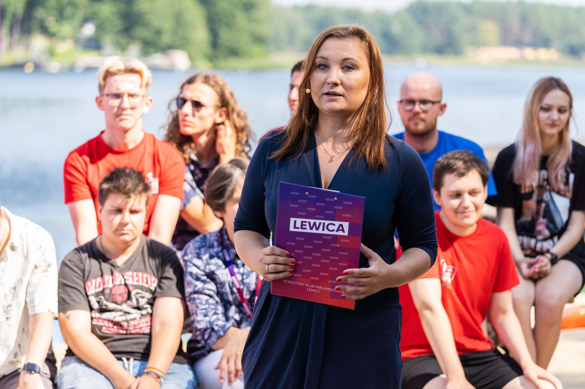 Kobieta w granatowej sukience przemawia podczas konwencji programowej Lewicy, trzymając fioletowy plakat z napisem LEWICA, a w tle siedzi grupa uczestników nad jeziorem.