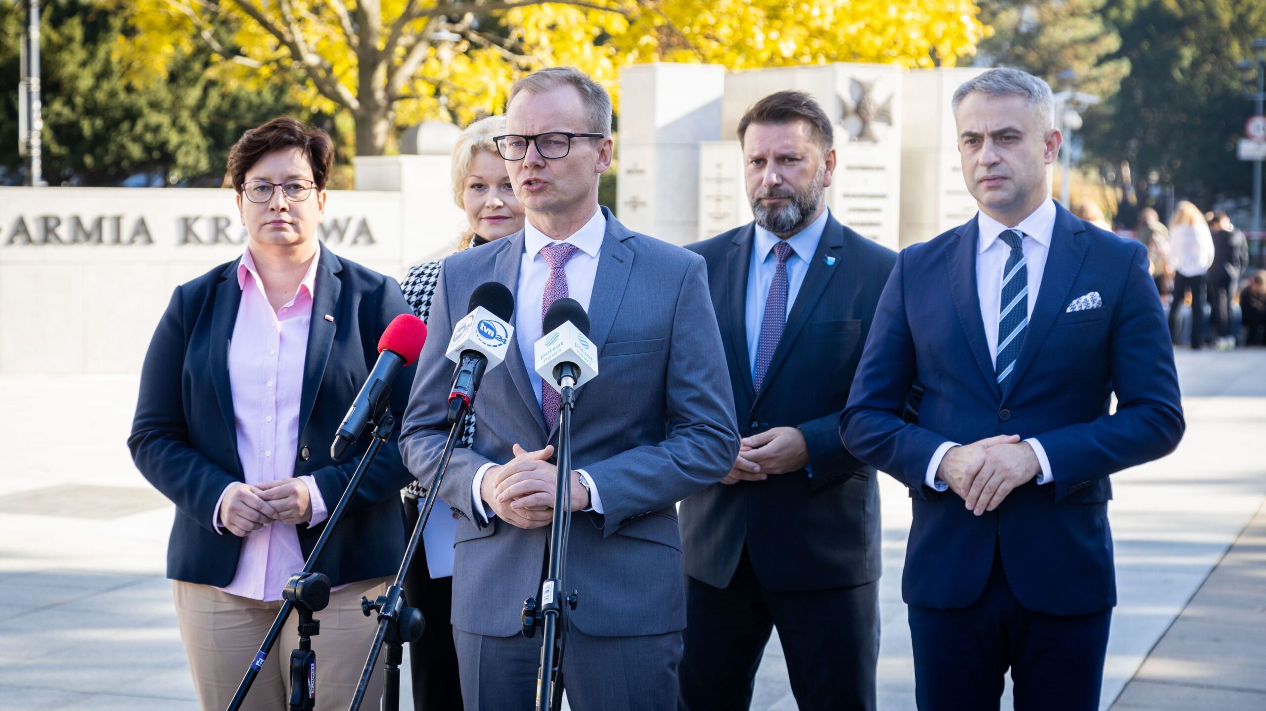 Na zdjęciu widocznych jest pięć osób w formalnych strojach, które stoją przed pomnikiem, podczas konferencji prasowej.