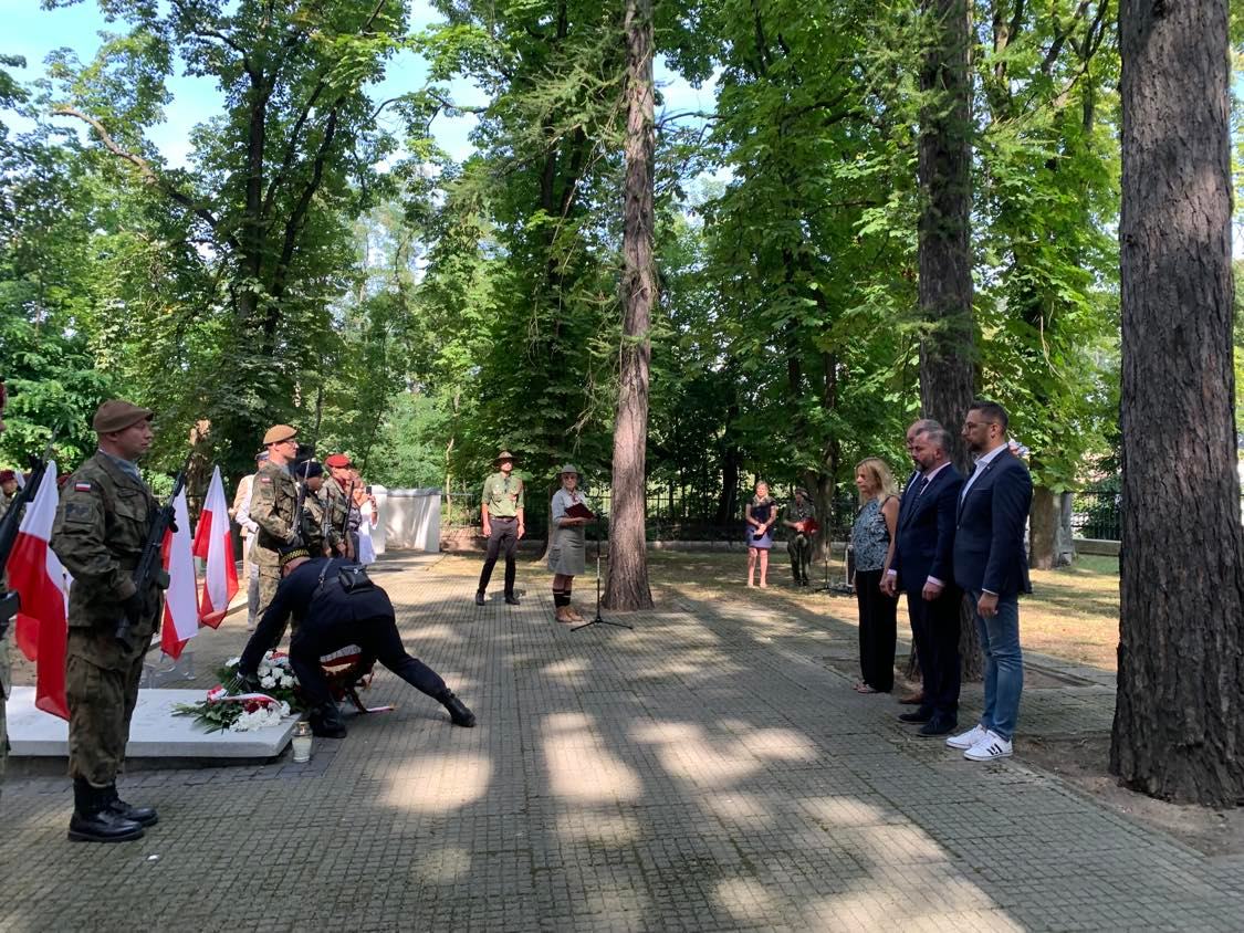 Na zdjęciu widać uroczystość związaną z składaniem wieńca, w której uczestniczą żołnierze oraz kilka osób w formalnym stroju, na tle zieleni drzew.