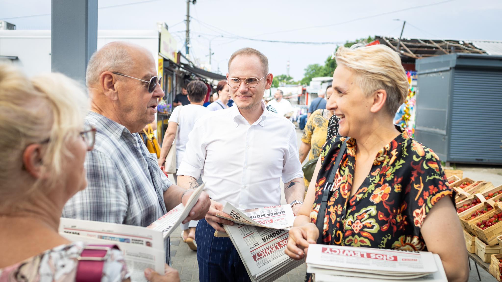 Politycy Lewicy w trasie rozmawiają z mieszkańcami na targowisku, rozdają gazety, a w tle widać stoiska z warzywami i owocami.