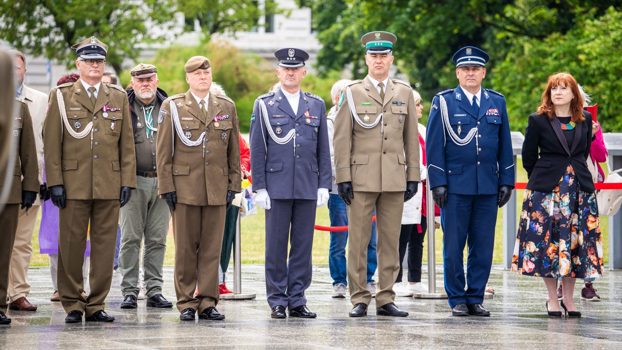 Grupa wojskowych i cywilów w mundurach oraz eleganckich strojach stoi na ceremonii na zewnątrz, w otoczeniu zieleni.