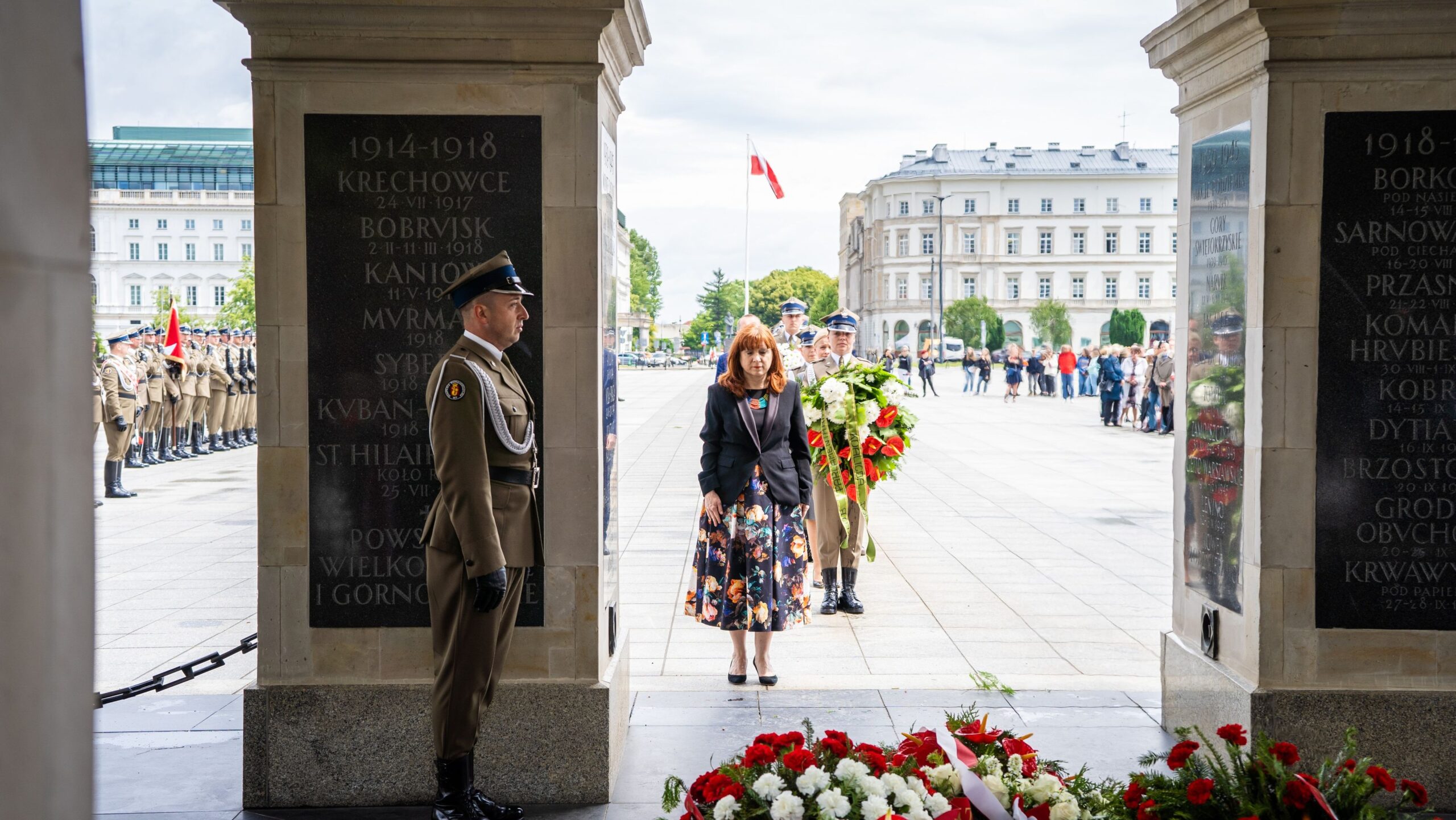 Na zdjęciu widoczna jest ceremonia złożenia kwiatów przez kobietę przy pomniku, otoczoną przez żołnierzy i z tłumem obserwatorów w tle.