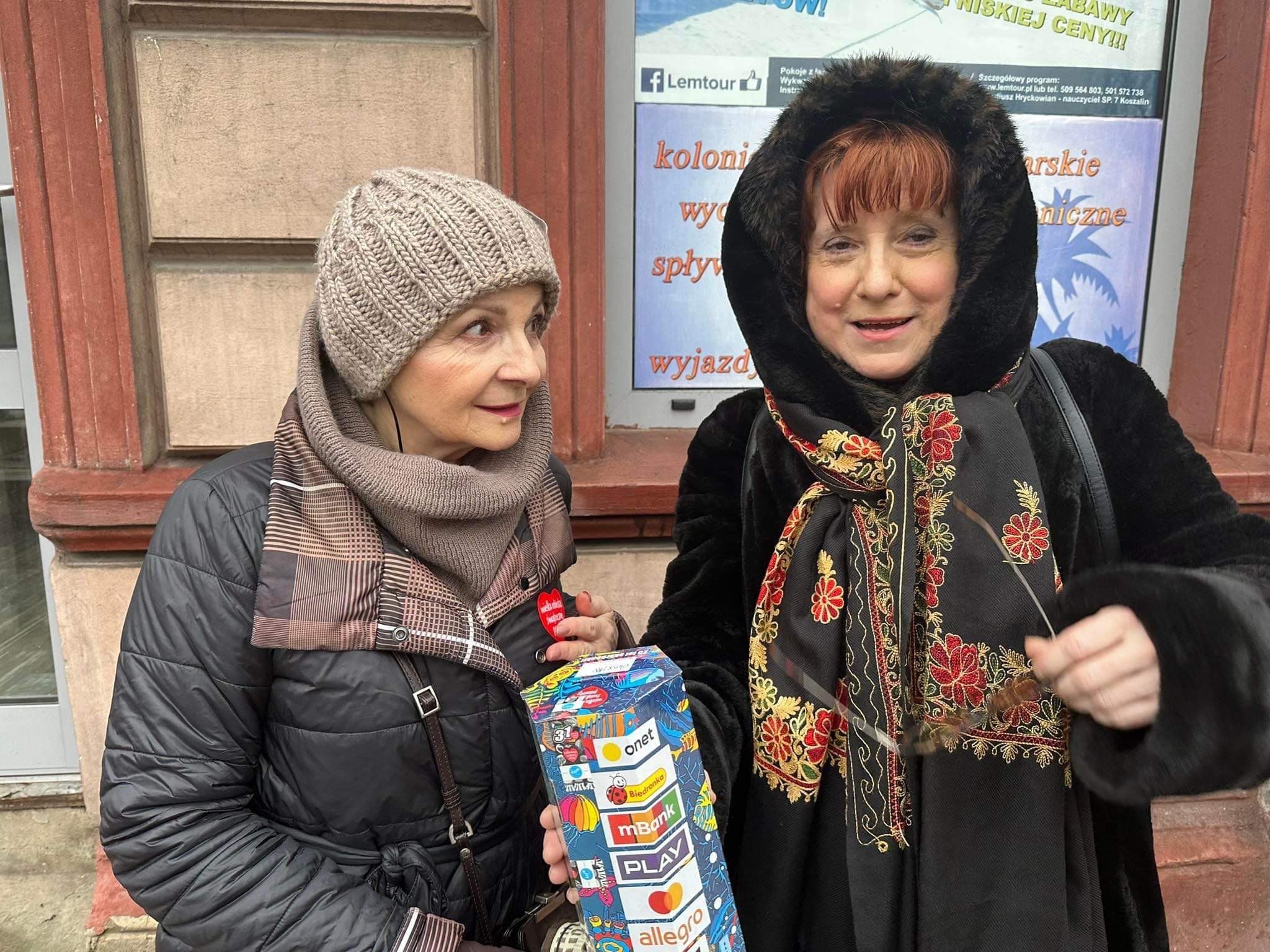 Na zdjęciu widać dwie kobiety stojące przed budynkiem, jedna z nich w czarnej futrzanej kurtce z kolorowym szalem, a druga w szarej czapce i kurtce.