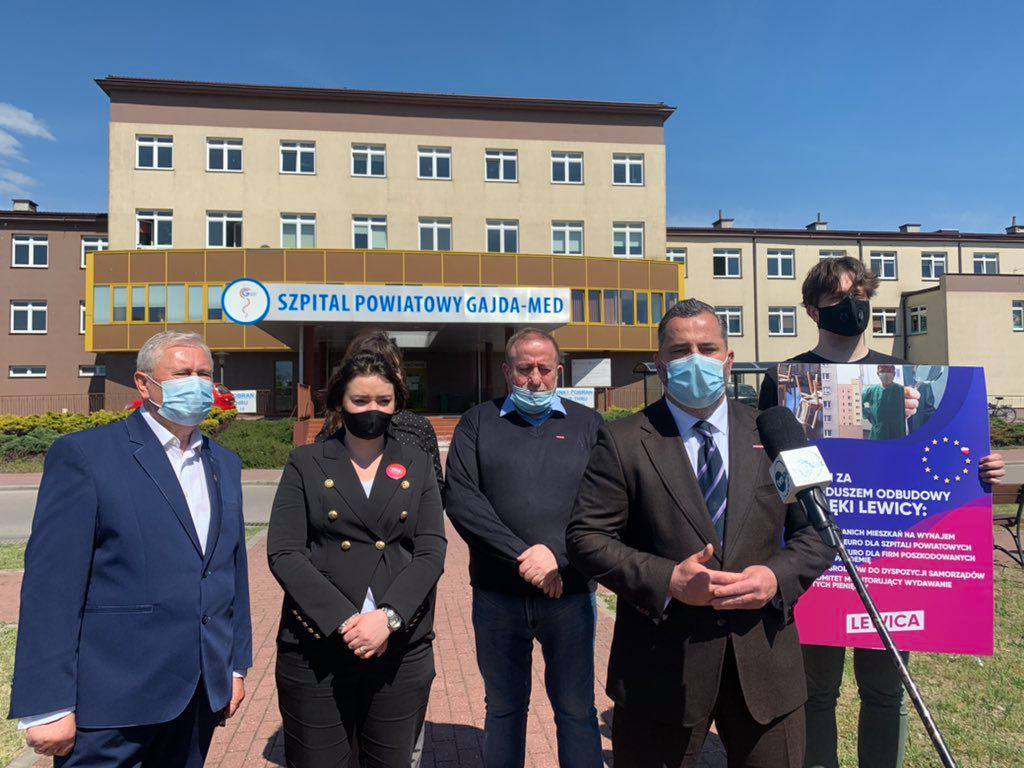 Pięć osób w maseczkach stoi przed wejściem do Szpitala Powiatowego Gajda-Med w Pułtusku, podczas konferencji prasowej o środkach z Funduszu Odbudowy, trzymając plakat Lewicy.