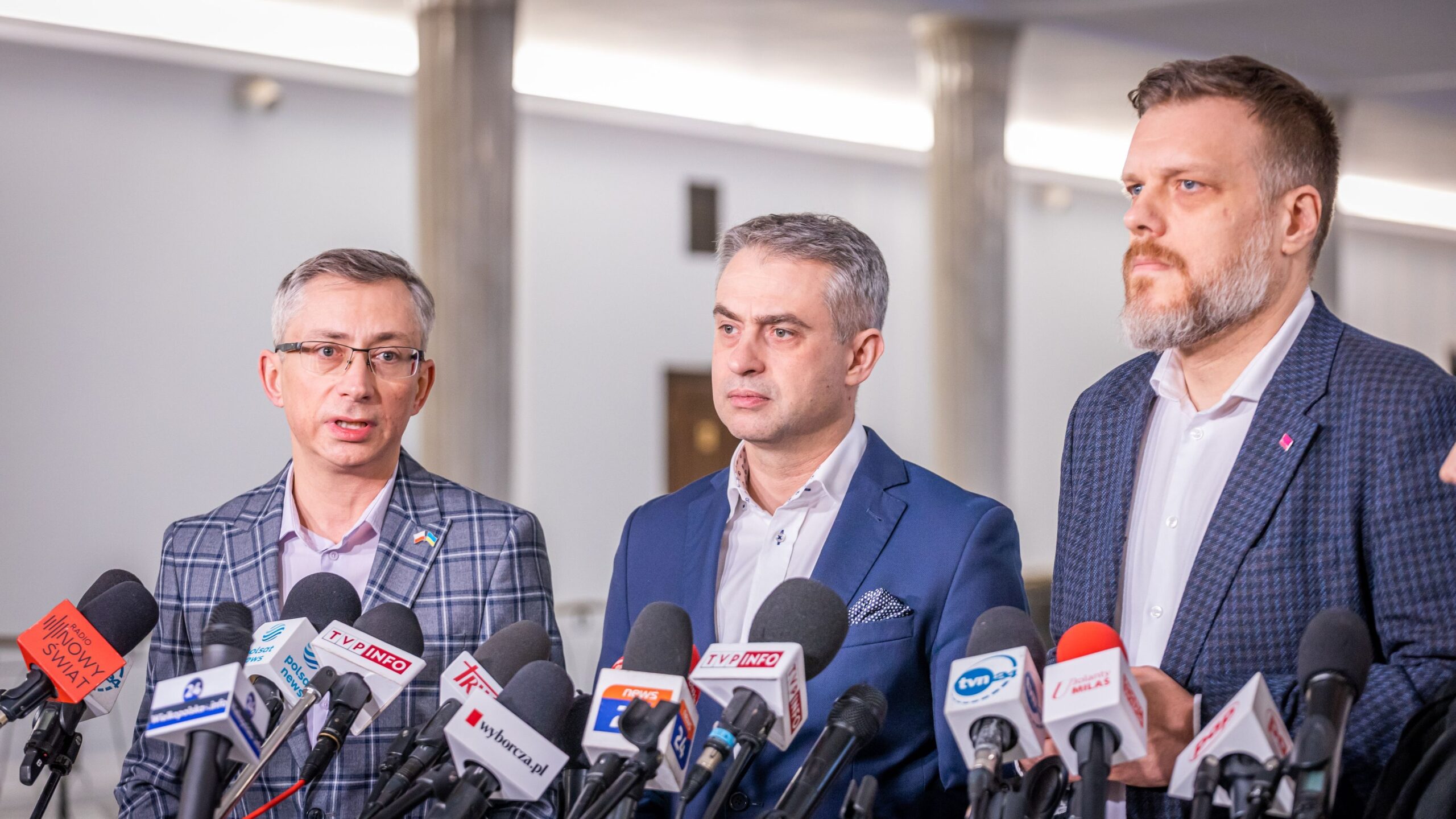 Na zdjęciu trzech mężczyzn w garniturach stoi przed mikrofonami i prowadzi konferencję prasową.