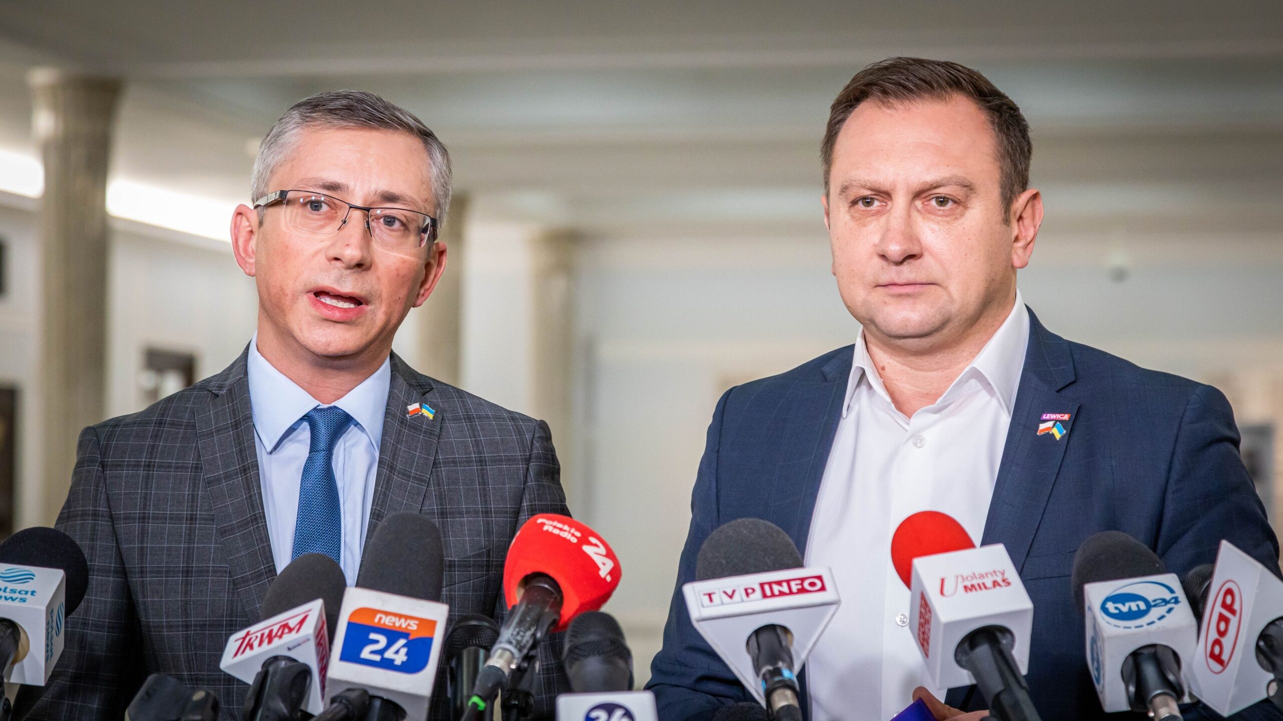 Dwóch mężczyzn w garniturach udziela konferencji prasowej, stojąc za licznymi mikrofonami przed mediami, w kontekście 2,7 mld zł dla TVP.