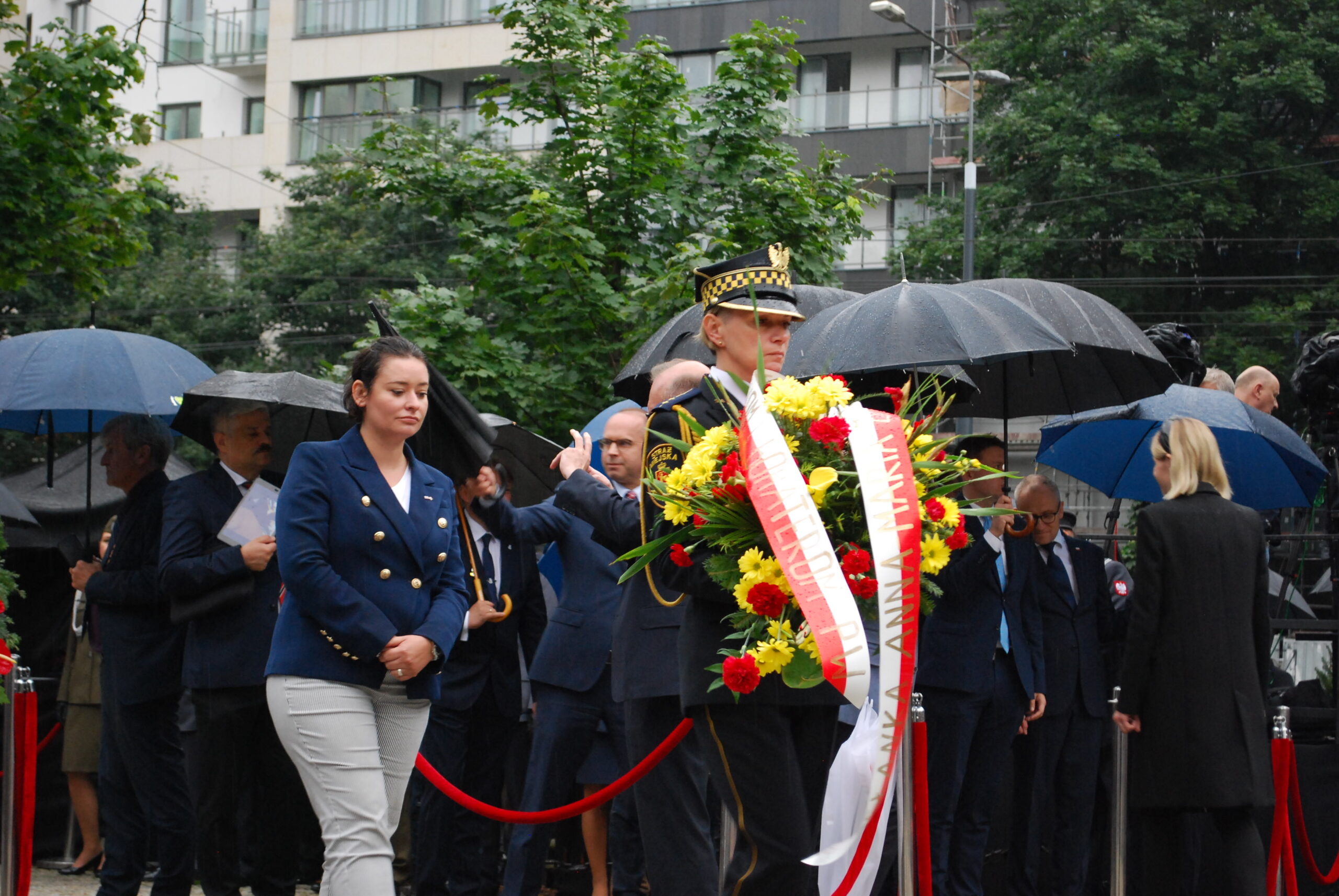 Uroczystość upamiętniająca ofiary Rzezi Woli w 77. rocznicę tragedii; funkcjonariusz w mundurze ceremonialnym niesie kwiatowy wieniec.