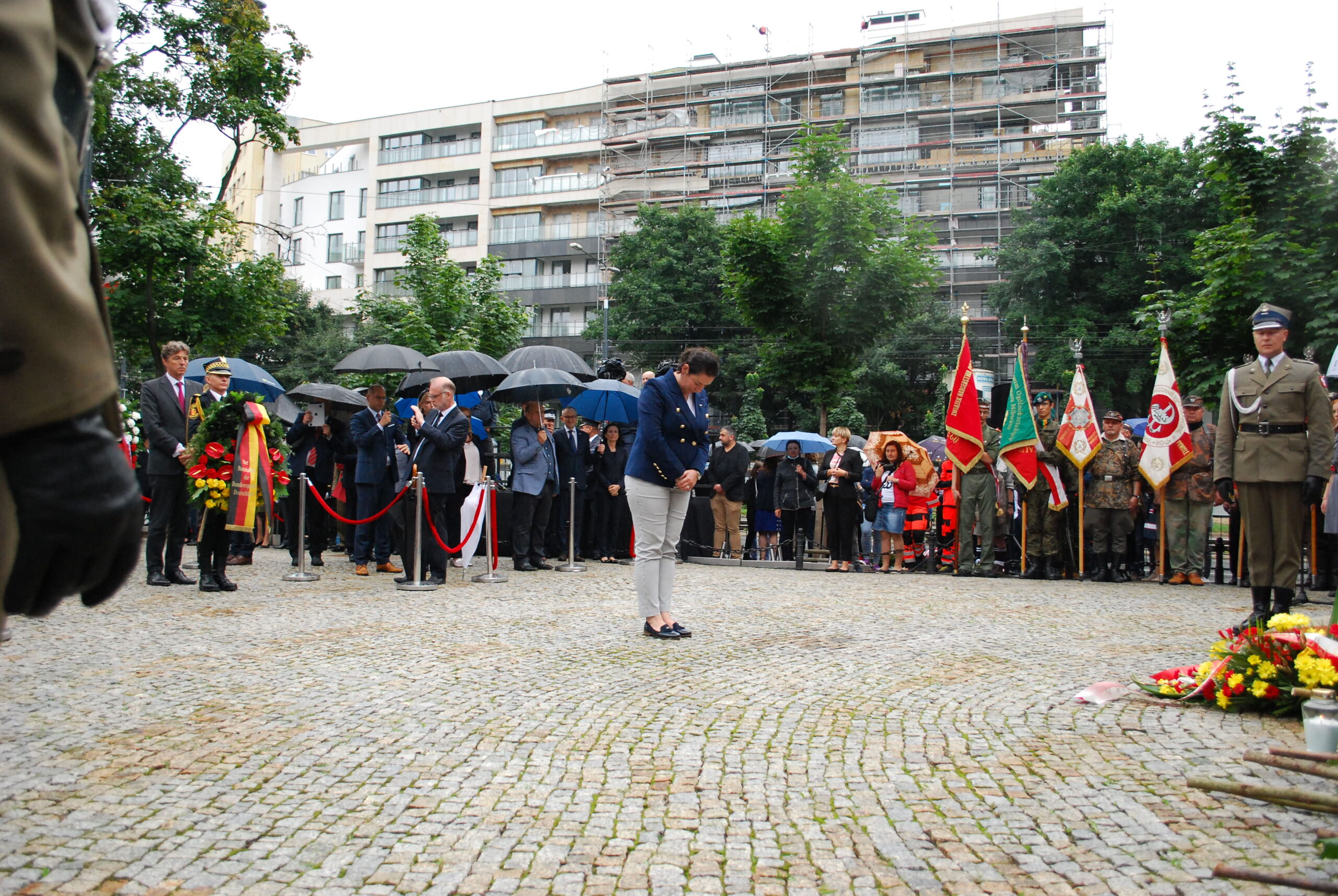 Uczestnicy ceremonii pamięci składają wieńce i oddają hołd ofiarom Rzezi Woli podczas 77. rocznicy tragedii, wśród zgromadzonych pod parasolami, z flagami i żołnierzami w mundurach.
