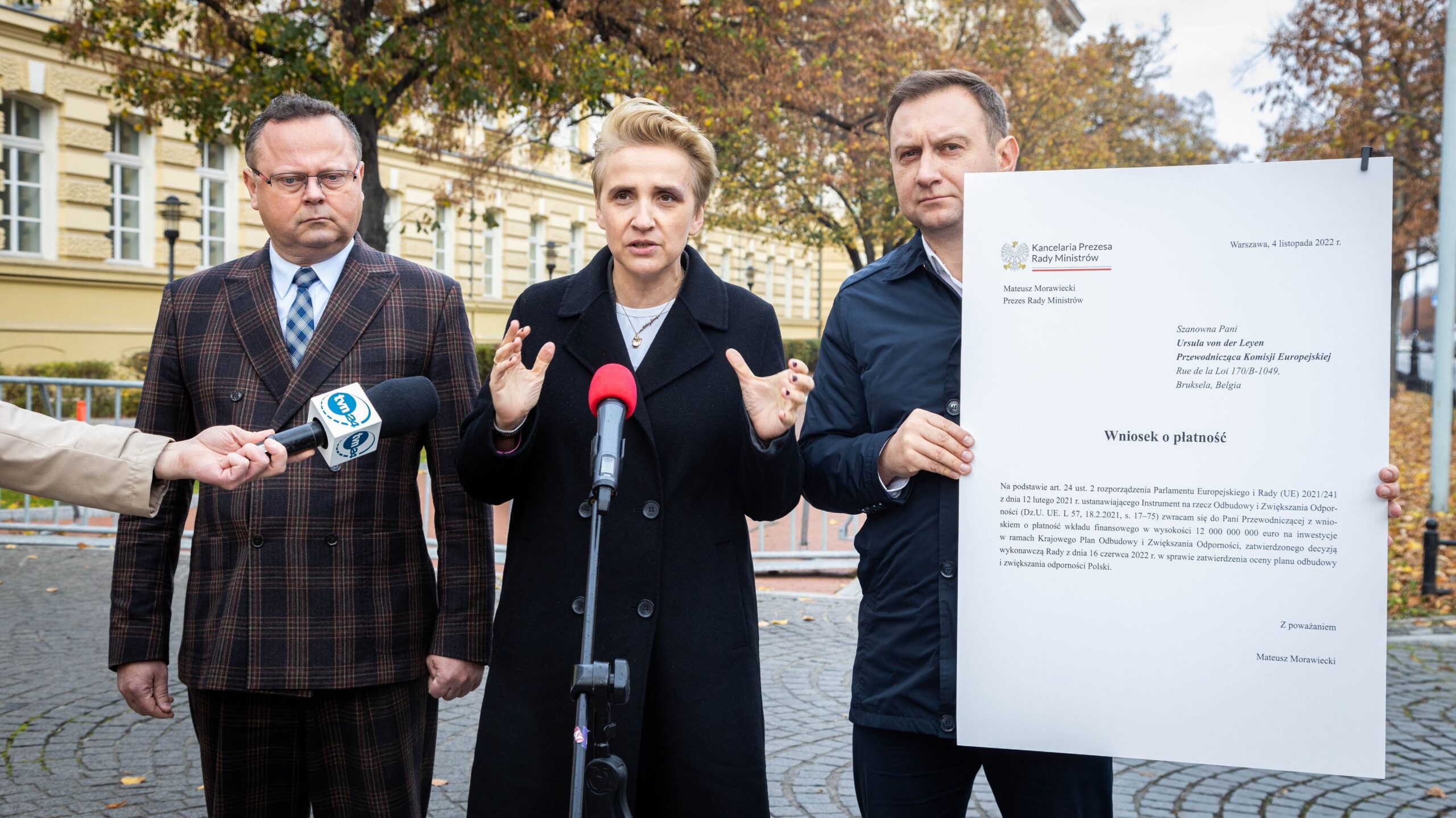 Na zdjęciu widać trzech polityków podczas konferencji prasowej, z jedną z osób mówiącą do mikrofonu oraz holding jednego z dokumentów.