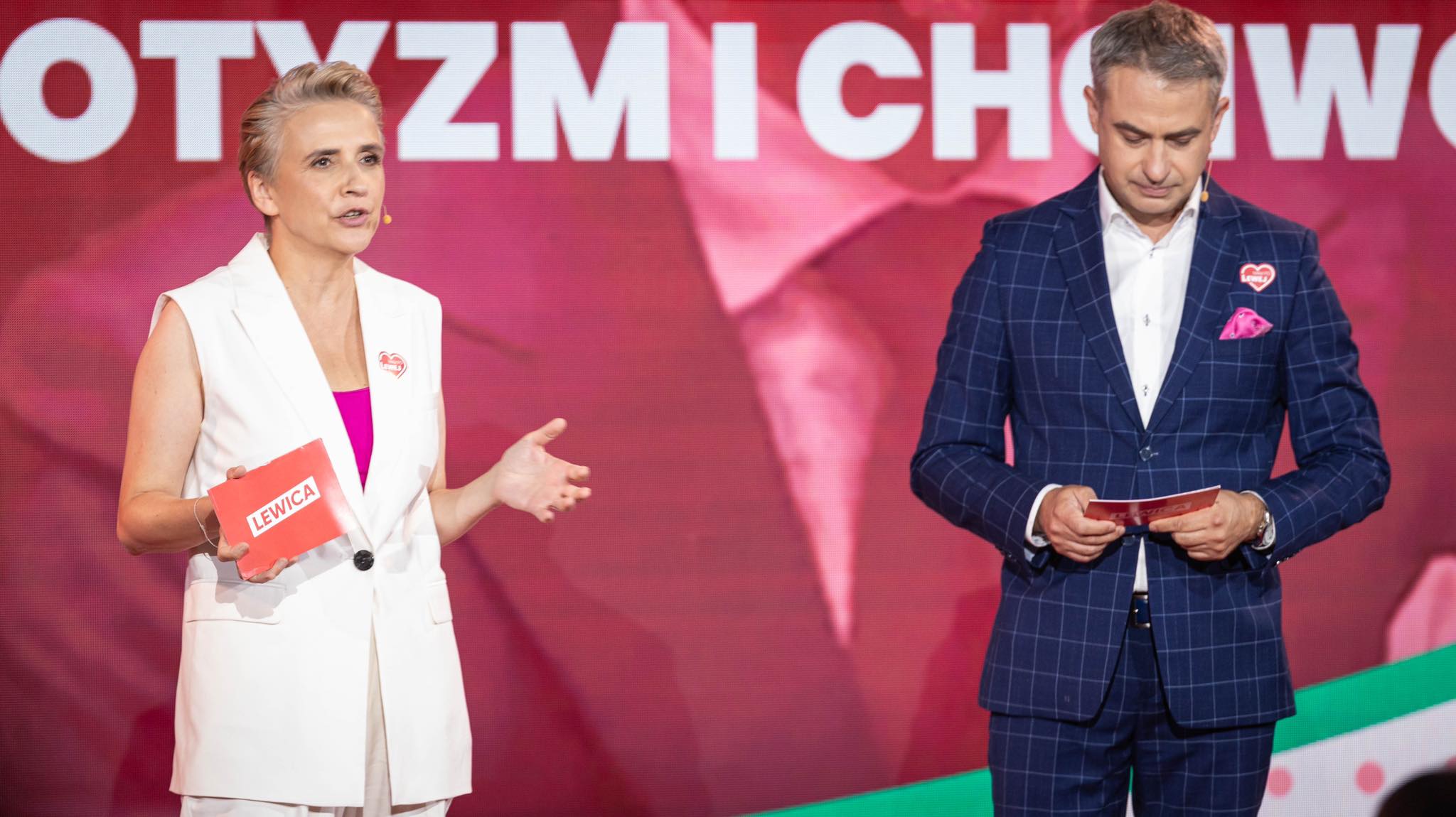 Na scenie dwie osoby prowadzące Lewicy; kobieta w białej kamizelce przemawia, trzymając kartkę z napisem Lewica, a mężczyzna w granatowym garniturze w kratę przegląda materiały, w tle widoczne hasło „Uczciwa praca musi popłacać”.