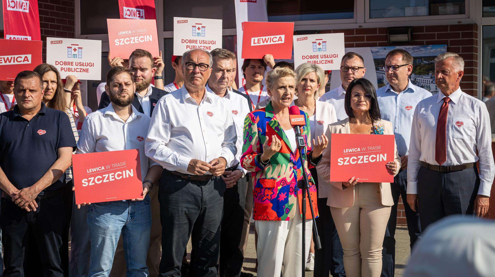 Grupa działaczy Lewicy w Szczecinie podczas konferencji, trzymają plakaty z napisem „SZCZECIN” i „Dobre usługi publiczne” oraz przemawia kobieta w kolorowej marynarce.