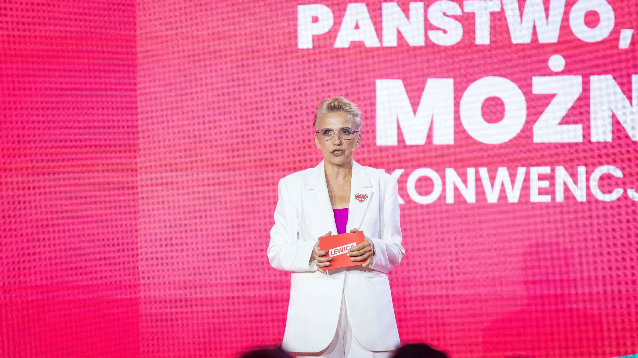 Kobieta w białym garniturze stoi na scenie na różowym tle z dużymi białymi napisami, trzyma czerwony notes z napisem LEWICA.