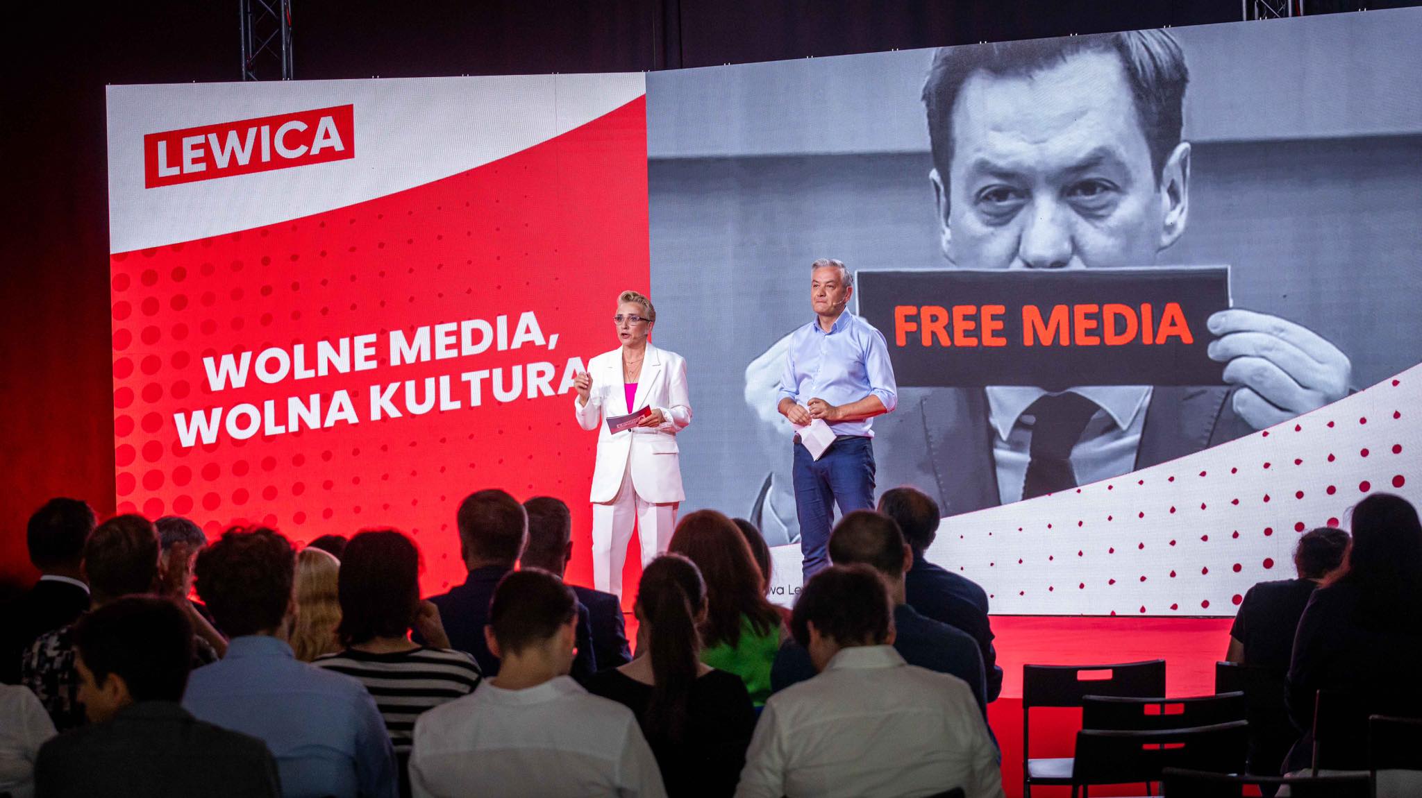 Para prowadzących przemawia na scenie podczas wydarzenia Lewicy, na tle dużego ekranu z czerwonym logo LEWICA i napisem „WOLNE MEDIA, WOLNA KULTURA”, obok czarno‑biały obraz z napisem „FREE MEDIA” przed zgromadzoną widownią.