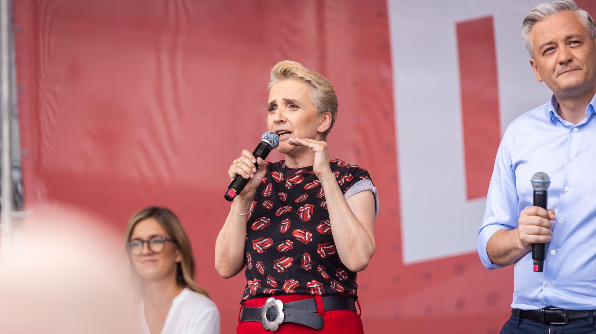 Kobieta na scenie przemawia do publiczności podczas wiecu w Częstochowie, trzyma mikrofon i gestykuluje, obok niej stoi mężczyzna w jasnej koszuli również trzymający mikrofon, na tle czerwonego baneru.