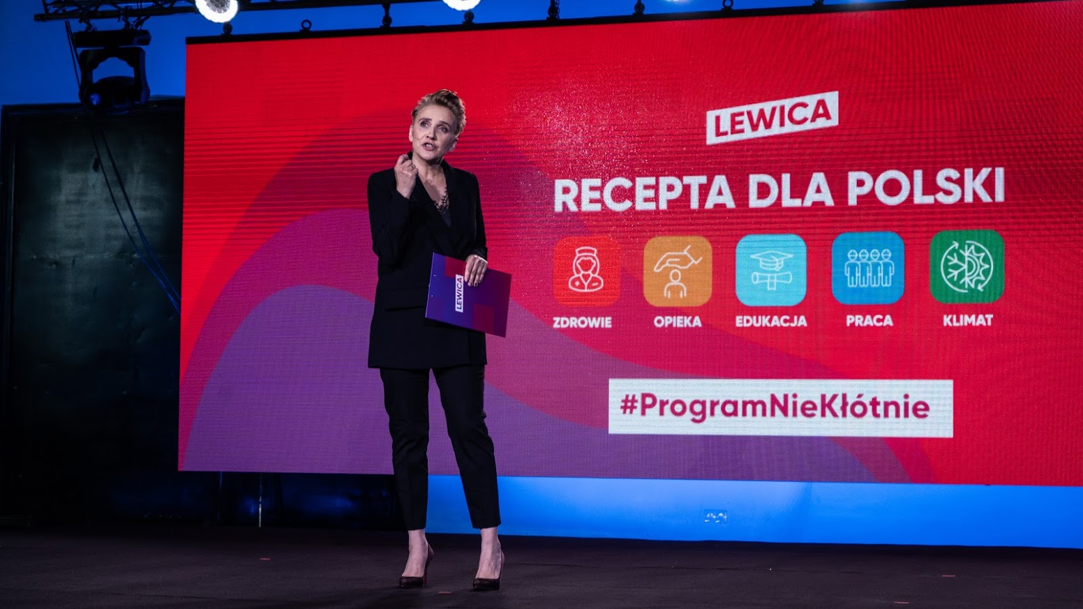 Kobieta w czarnym garniturze stoi na scenie, trzyma notatnik, a w tle widoczny jest czerwony baner Lewica z napisem „Recepta dla Polski” i ikonami zdrowia, opieki, edukacji, pracy oraz klimatu.