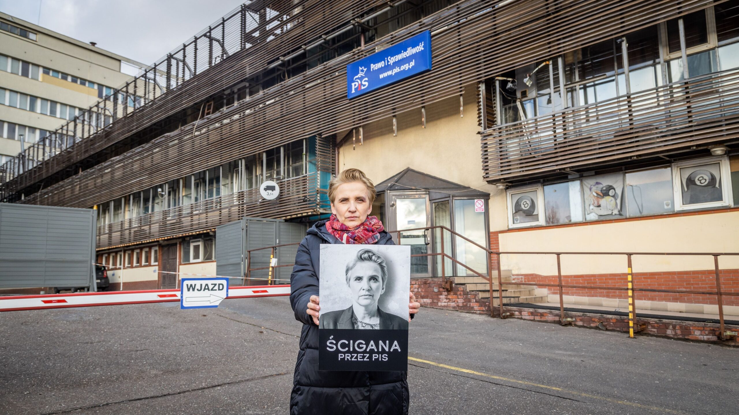 Na zdjęciu widać kobietę stojącą przed budynkiem z siedzibą partii politycznej, trzymającą plakat z napisem 