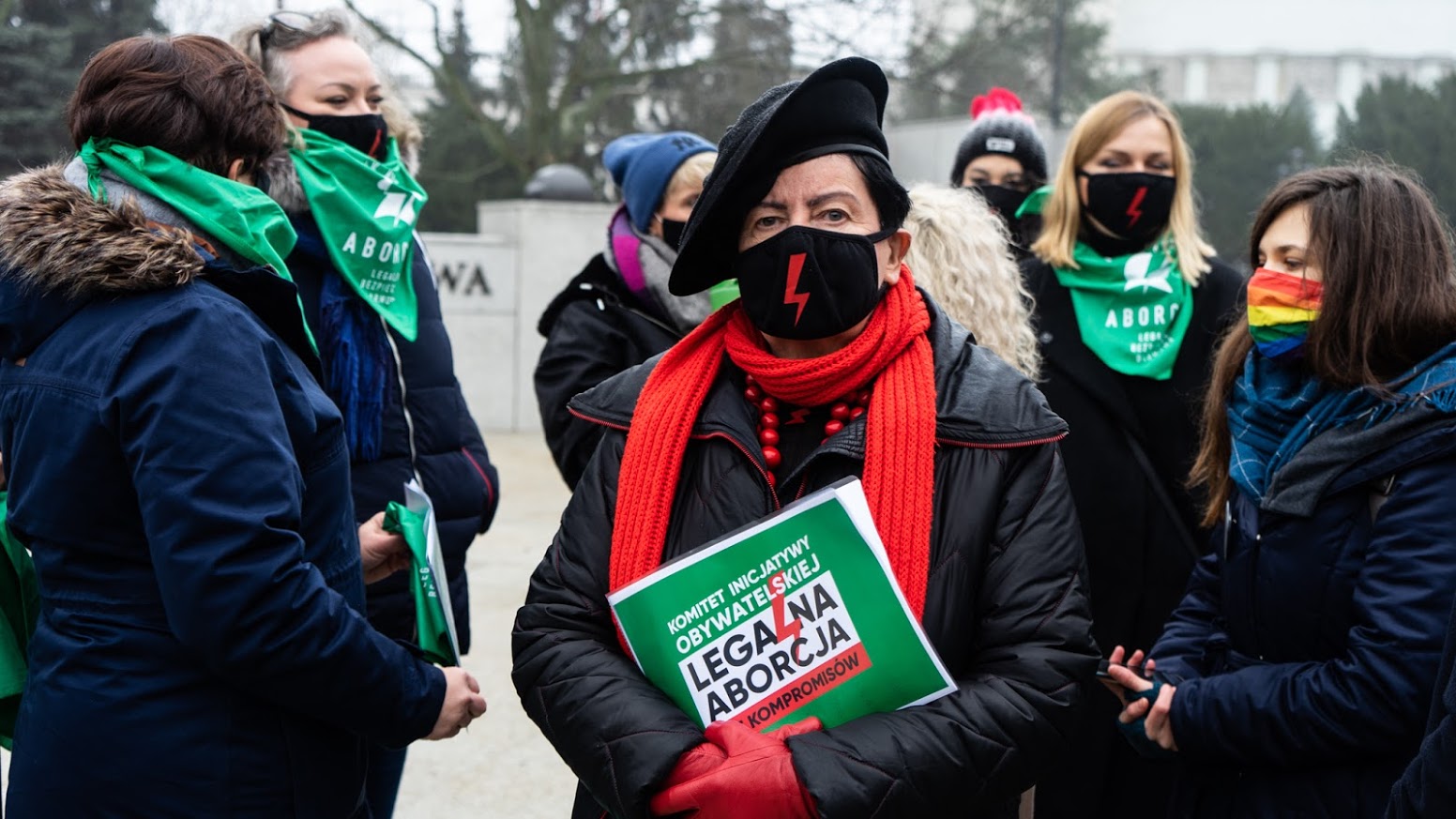 Grupa demonstrantów popierających projekt „Legalna Aborcja. Bez Kompromisów”; na pierwszym planie kobieta w czarnej kurtce z czerwonym szalem i maseczką z czerwonym symbolem trzyma zieloną tablicę z napisem „KOMITET INICJATYWY OBYWATELSKIEJ LEGALNA ABORCJA KOMPROMISÓW”.