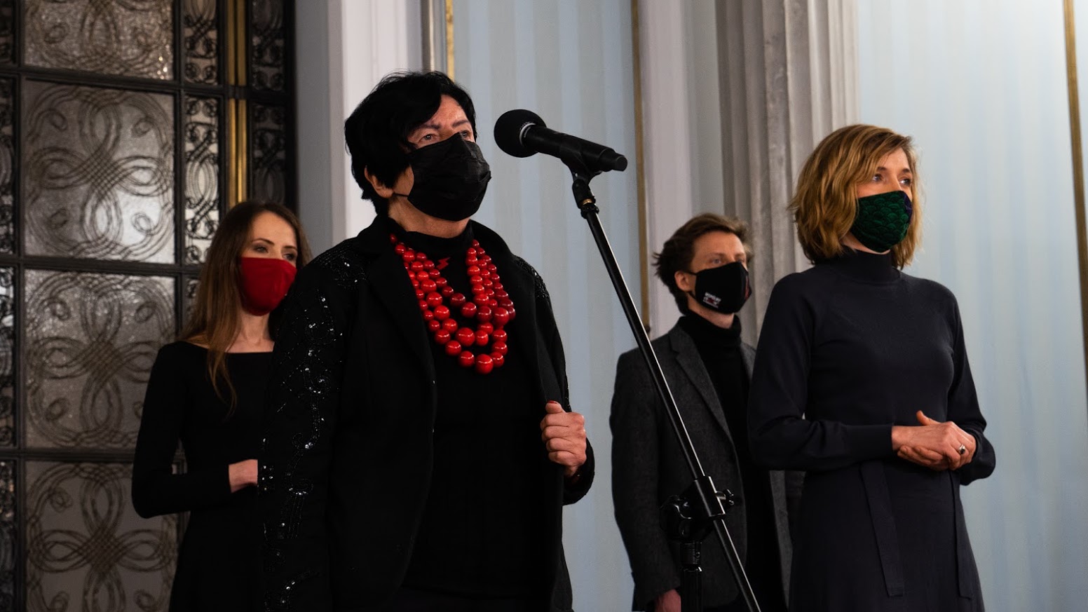 Grupa osób w maseczkach stoi przy mikrofonie w eleganckim wnętrzu; na pierwszym planie po lewej kobieta nosząca duży czerwony naszyjnik z koralików, po prawej druga kobieta w ciemnym stroju.