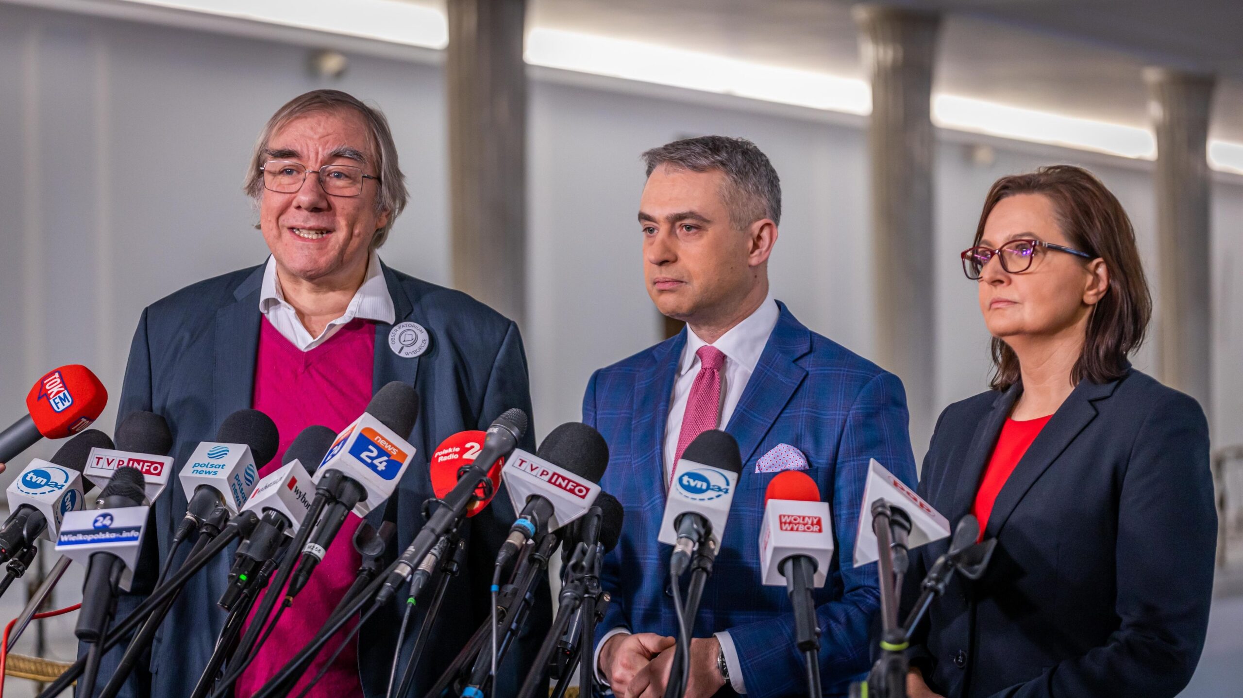Trzy osoby stoją przed mikrofonami na konferencji prasowej, każda w eleganckim stroju, w tle widać jasne wnętrze budynku.