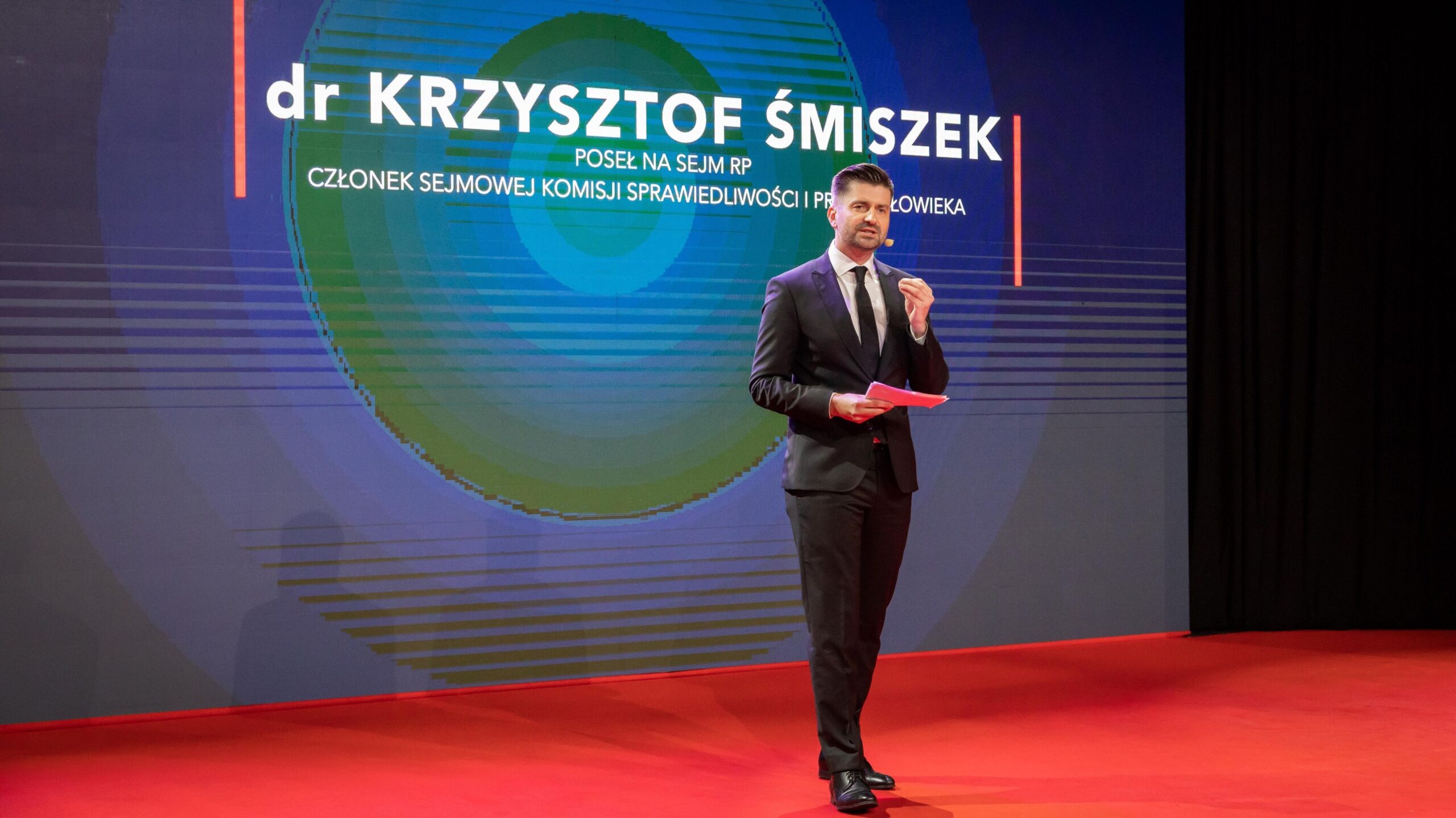 Mężczyzna w eleganckim garniturze przemawia podczas wydarzenia, z tłem wskazującym jego imię i nazwisko oraz pełnioną funkcję w parlamencie.