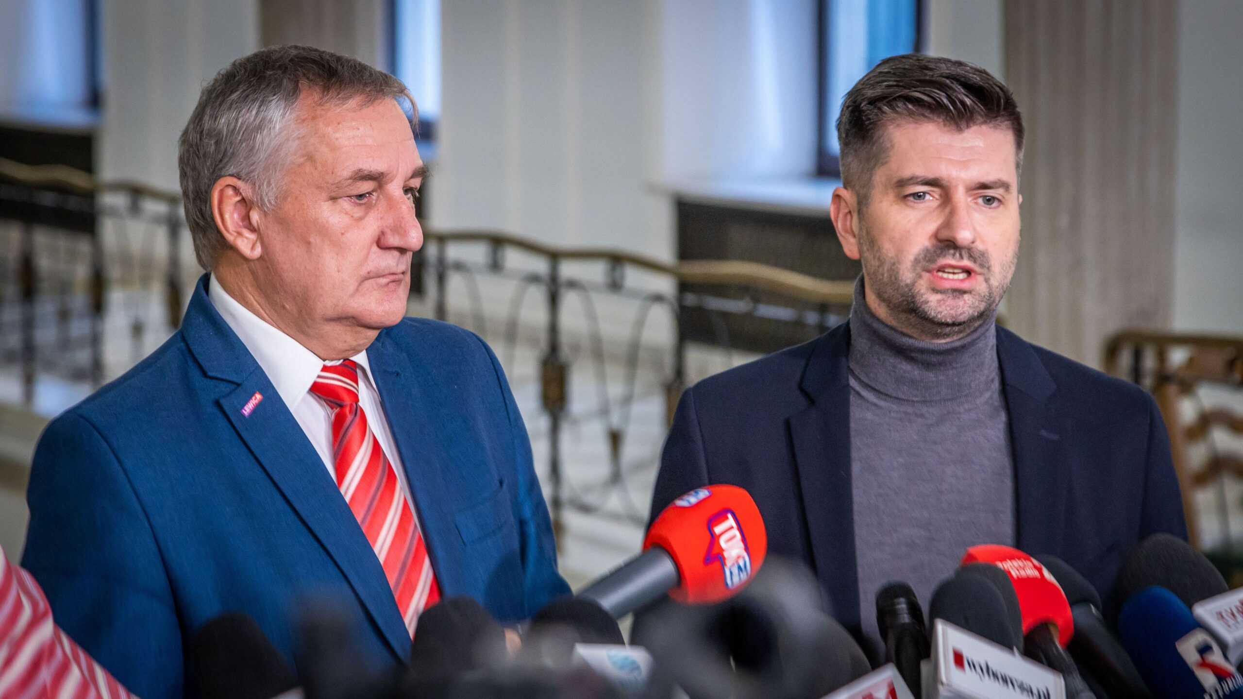 Dwóch mężczyzn w garniturach prowadzi konferencję prasową, stojąc przed licznymi mikrofonami w wnętrzu z balustradą w tle.