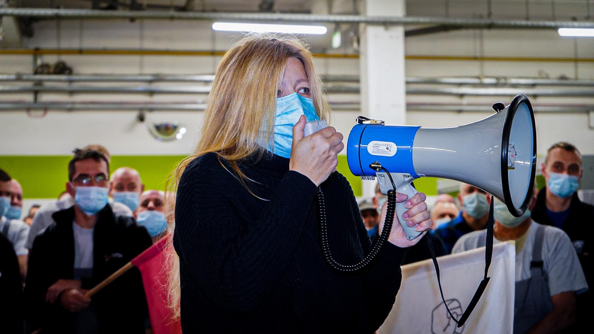 Na zdjęciu kobieta w maseczce trzyma megafon, przemawiając do zgromadzonego tłumu, z mężczyznami w tle, również w maseczkach.