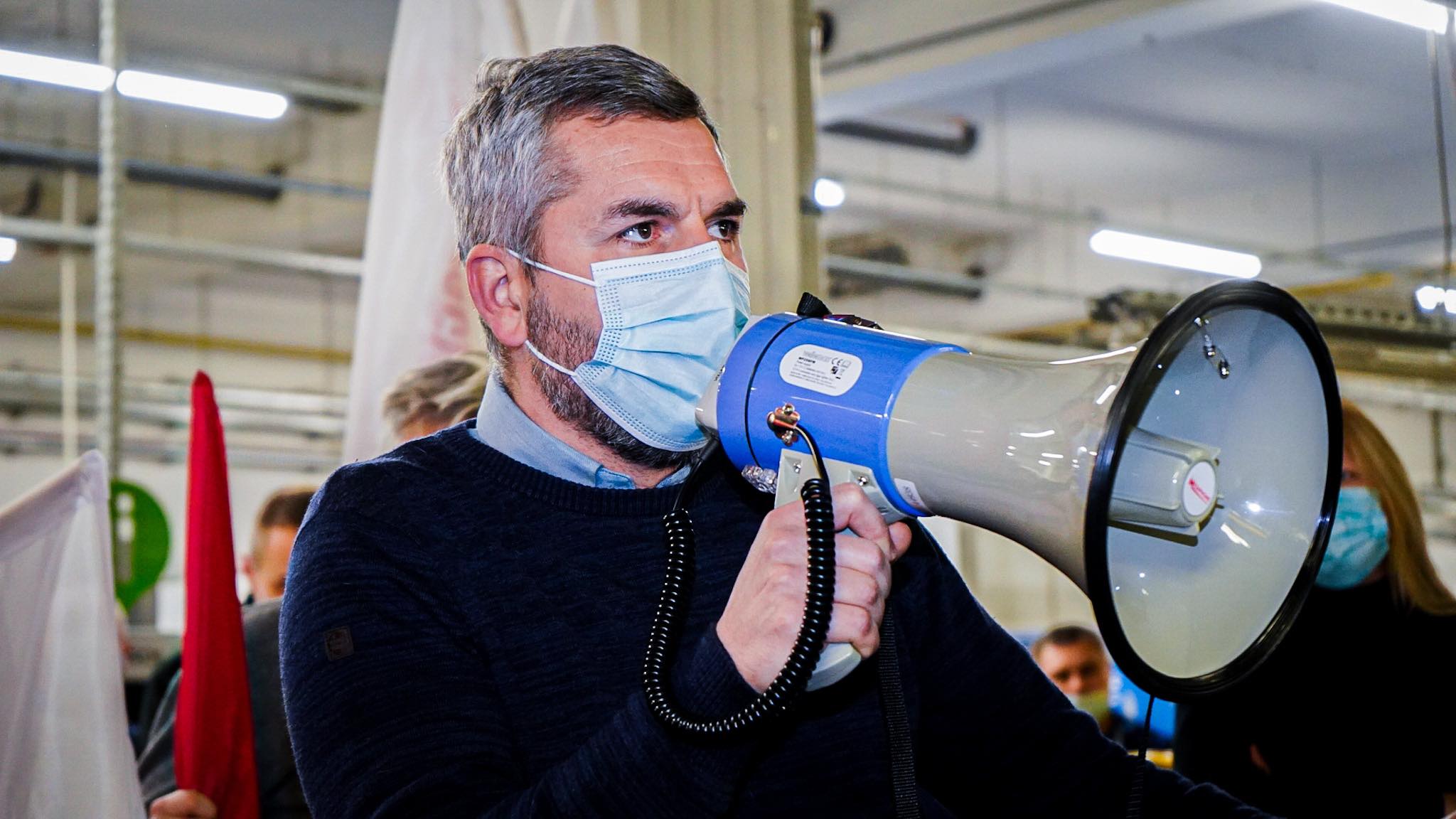 Na zdjęciu widać mężczyznę w niebieskiej maseczce, trzymającego megafon, który przemawia do grupy ludzi w tle.