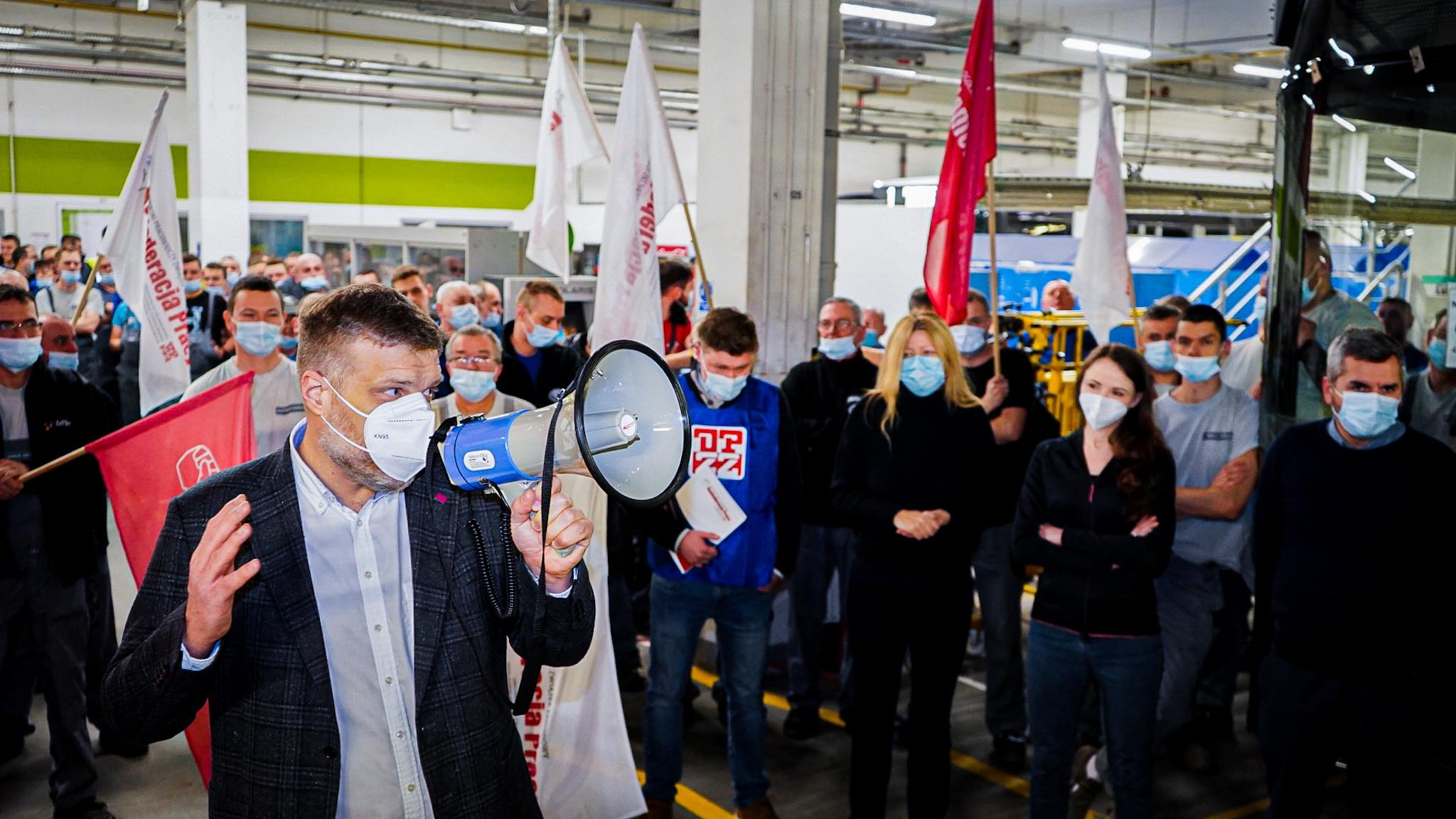 Pracownik przemysłowy przemawia do grupy ludzi w maseczkach, trzymając megafon, podczas zgromadzenia w zakładzie produkcyjnym.