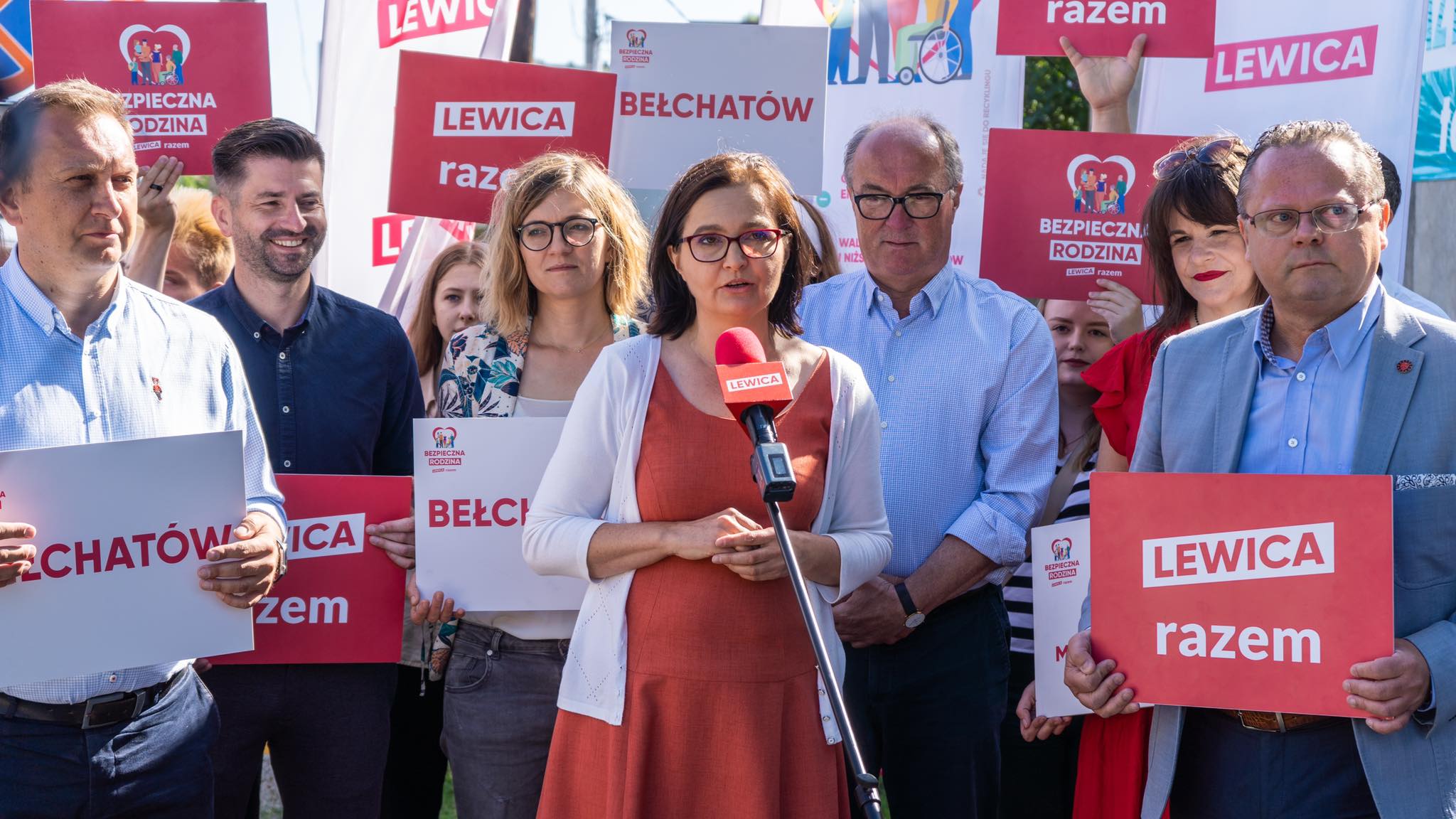 Grupa ludzi na plenerowym wiecu politycznym z plakatami „LEWICA razem”, a w centrum kobieta w pomarańczowej sukience przemawia do mikrofonu.