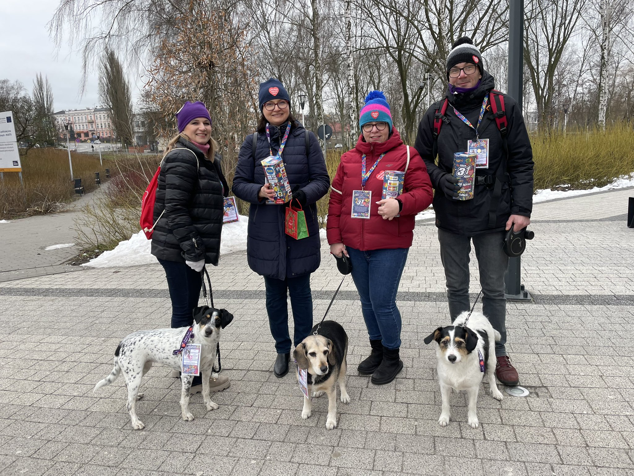 Cztery osoby w zimowych kurtkach stoją na chodniku z trzema psami, mają identyfikatory WOŚP 2023 i gadżety zbiórkowe.