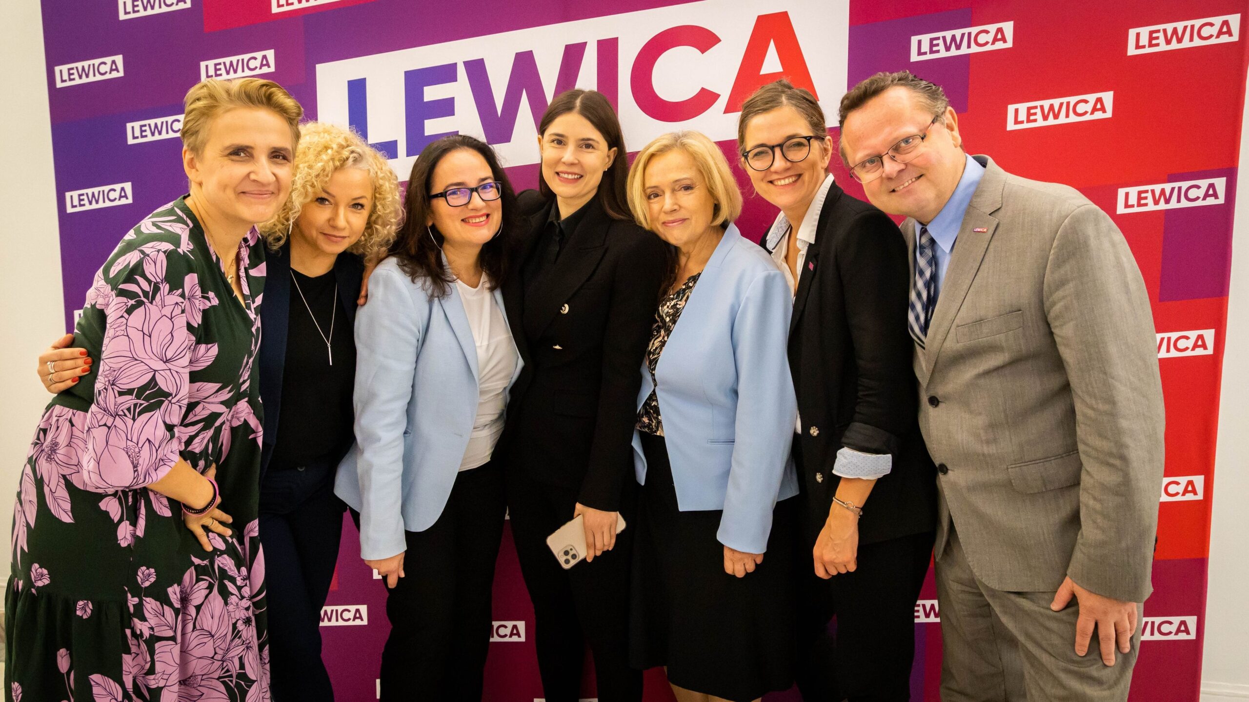 Na zdjęciu grupa osób pozująca wspólnie przed tłem z logo partii Lewica.