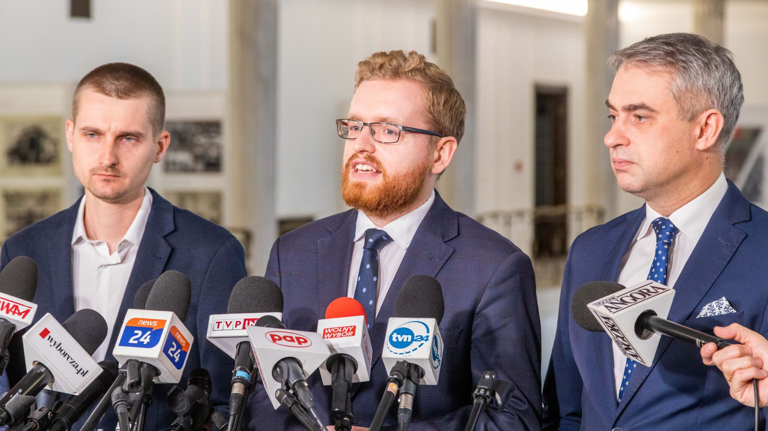 Trzech mężczyzn w garniturach stoi przed mikrofonami prasowymi podczas konferencji prasowej.