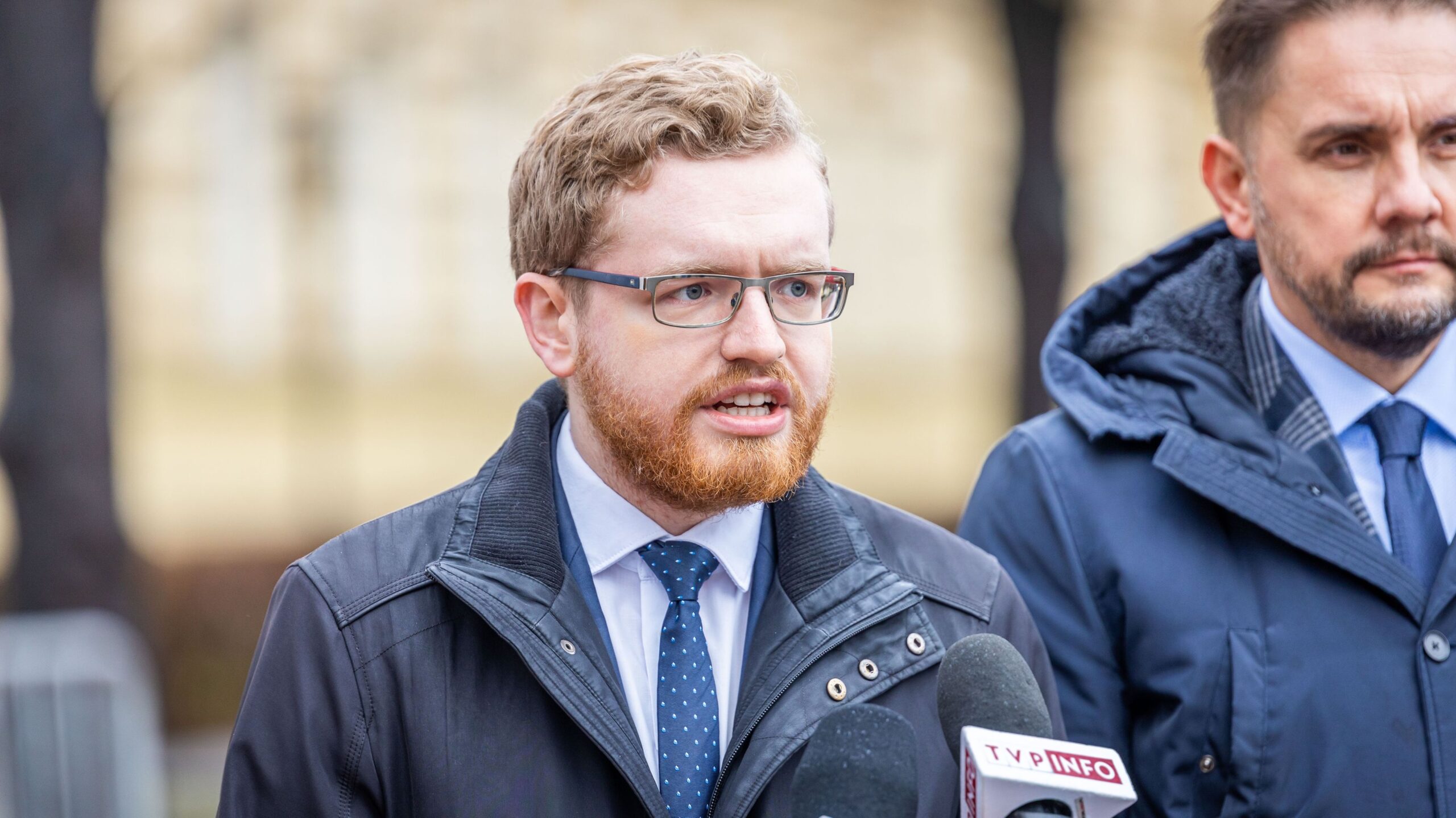 Mężczyzna z czerwonym zarostem i okularami przemawia do mediów przy mikrofonach podczas konferencji prasowej o inflacji, stojąc obok kolegi w marynarce.