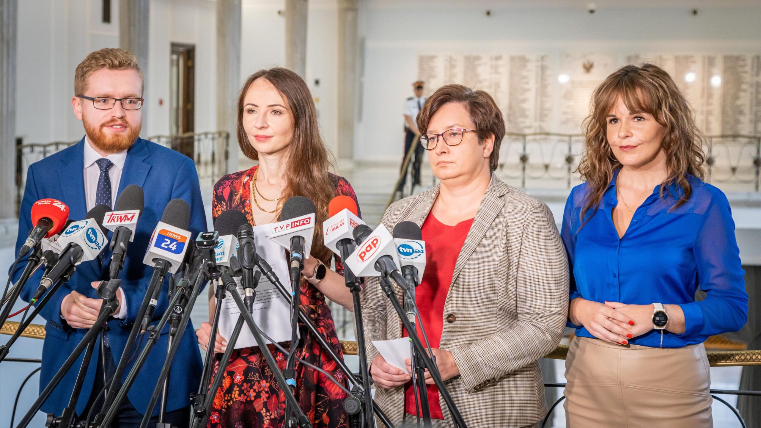 Na zdjęciu widoczni są czterej mówcy stojący przed mikrofonami podczas konferencji prasowej w eleganckim wnętrzu.