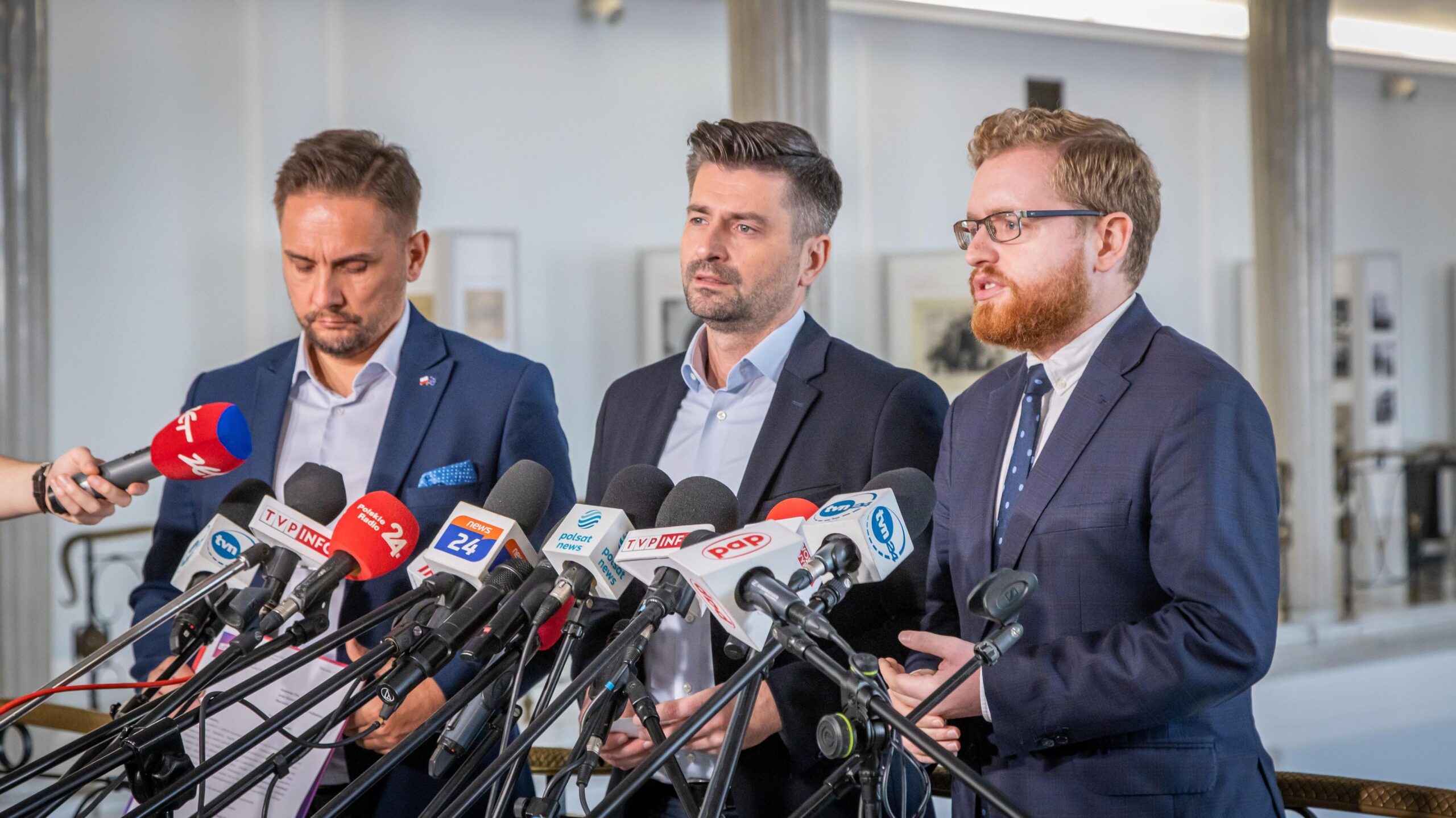 Na zdjęciu trzej mężczyźni w garniturach stoją przed mikrofonami i prowadzą konferencję prasową.