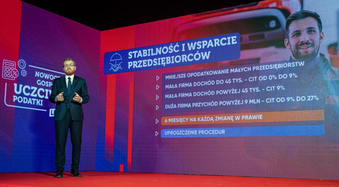 Na zdjęciu prezentuje się mężczyzna w garniturze, przemawiający na tle ekranu z informacjami na temat stabilności i wsparcia dla przedsiębiorców.
