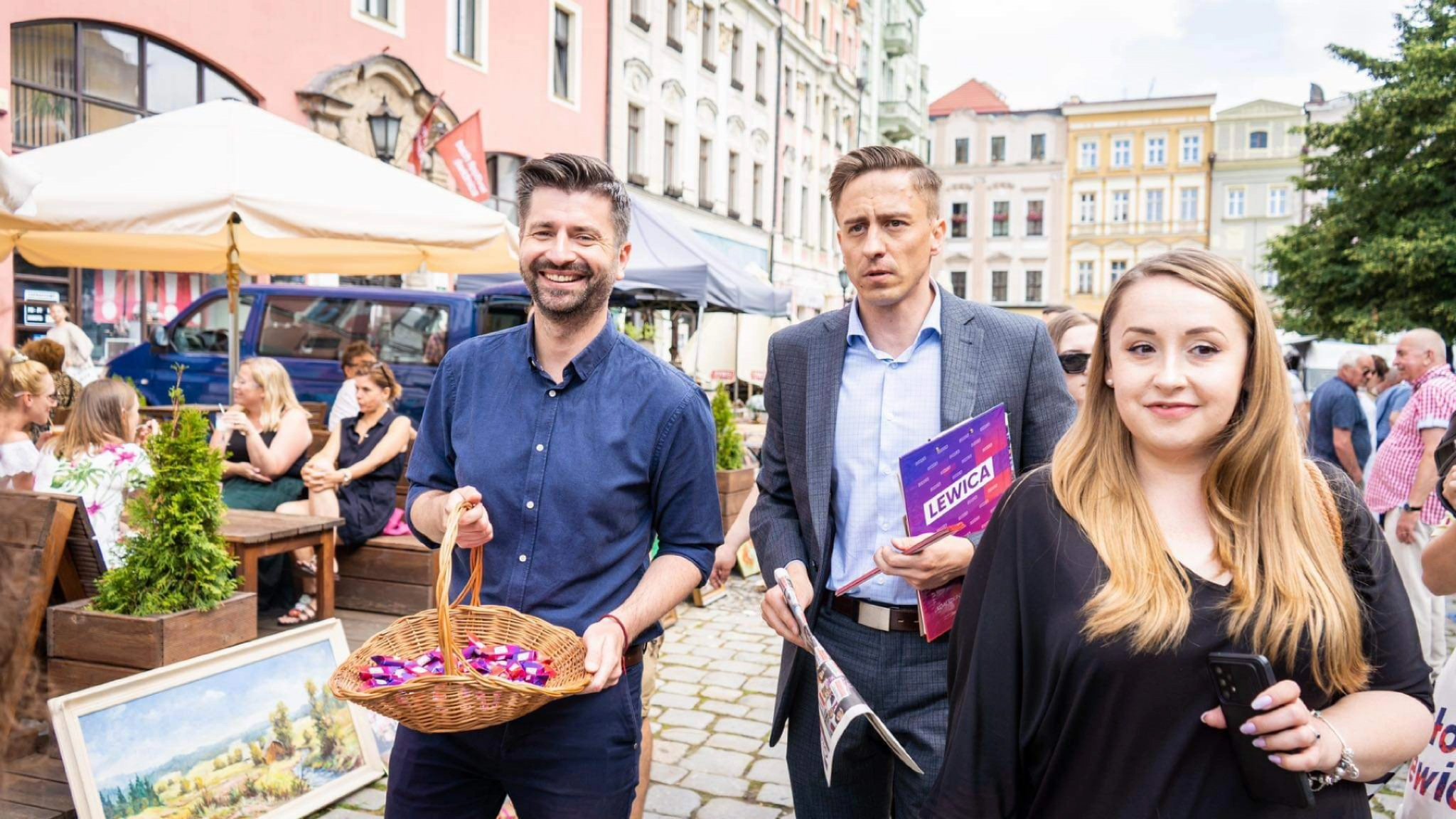 Trzy osoby spacerują po świdnickim rynku podczas wydarzenia Lewicy w trasie: mężczyzna w granatowej koszuli niesie kosz z cukierkami, drugi w garniturze trzyma ulotki, a kobieta obok trzyma telefon.
