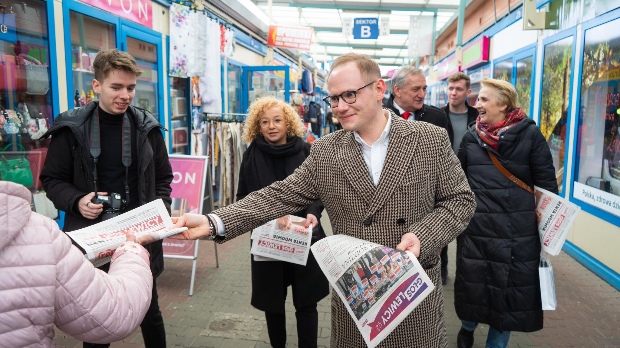 Grupa osób w przejściu handlowym w Szczecinie, a wśród nich mężczyzna w kraciastym płaszczu rozdaje gazetę przechodniom.