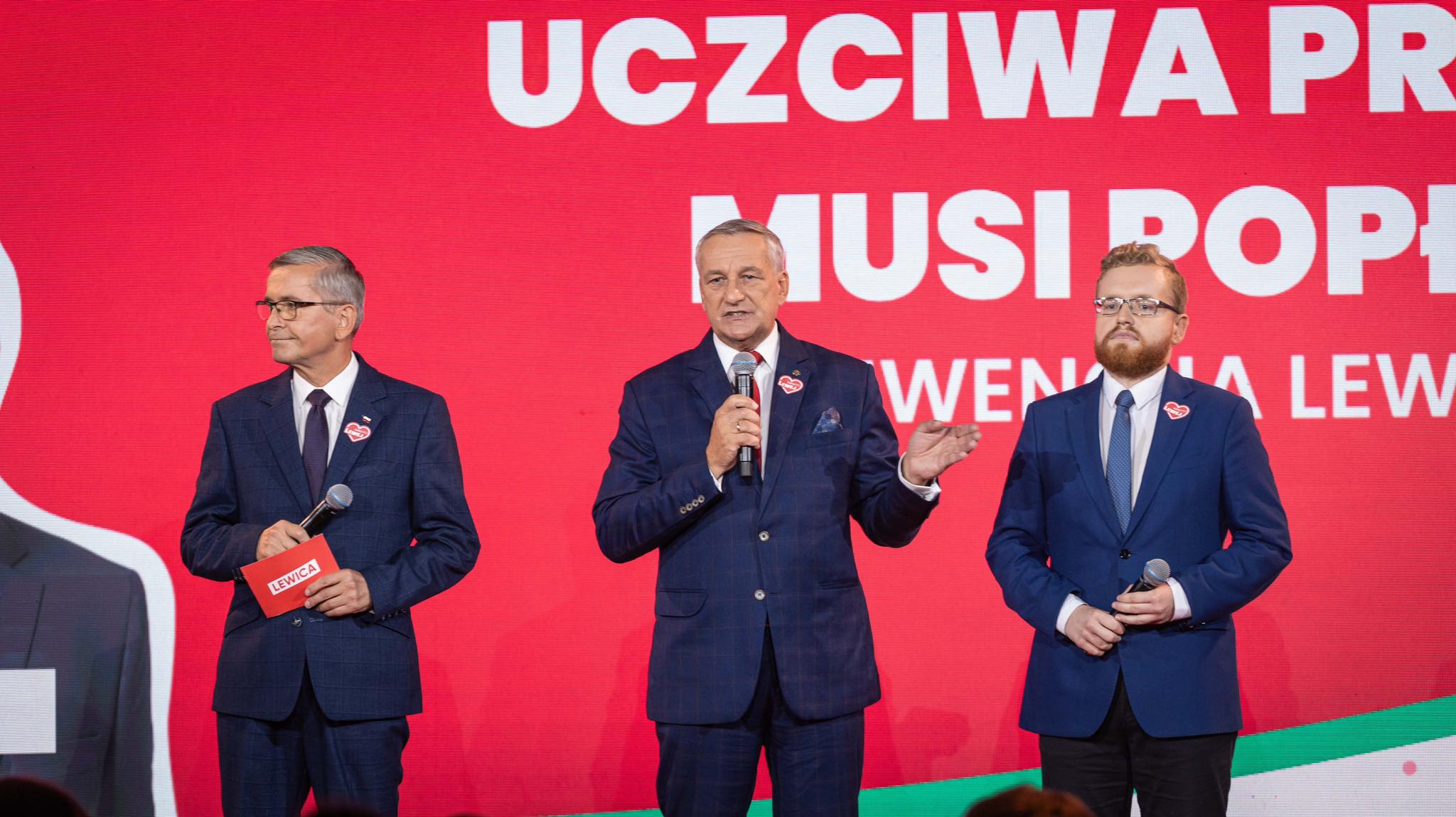 Trzech mężczyzn w garniturach stoi na scenie na czerwonym tle z hasłem „Uczciwa praca musi popłacać”; jeden z nich przemawia do mikrofonu.