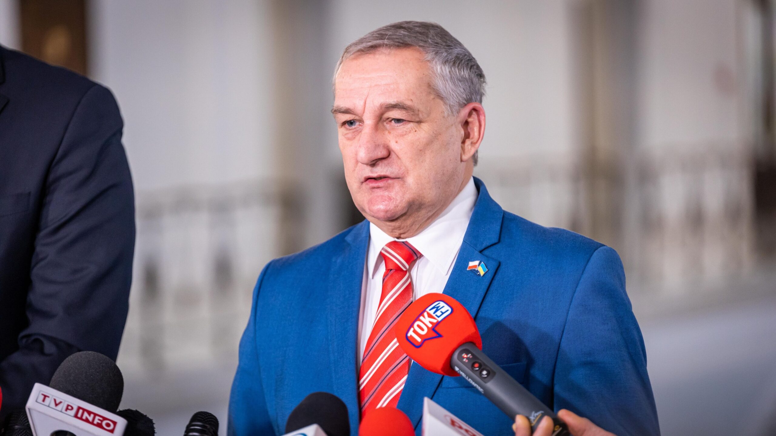 Na zdjęciu widoczny jest mężczyzna w niebieskim garniturze z czerwonym krawatem, który udziela wywiadu dziennikarzom otoczony mikrofonami.