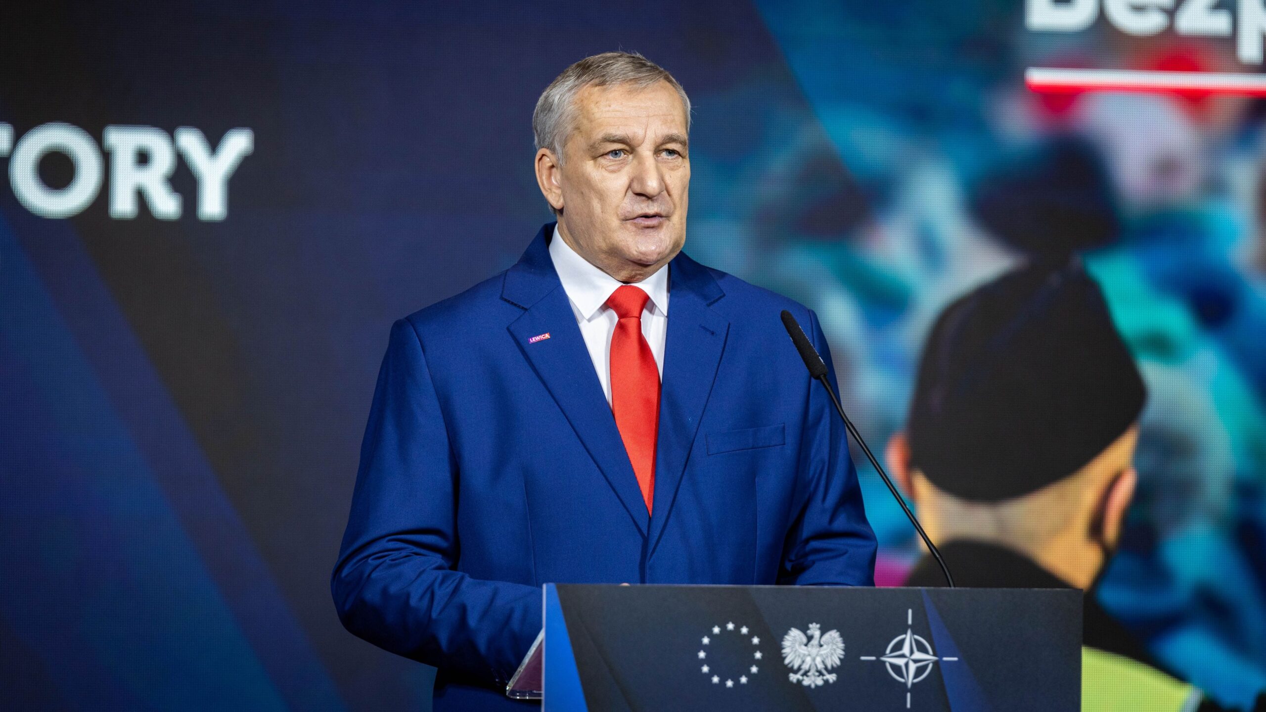 Mężczyzna w granatowym garniturze i czerwonym krawacie przemawia z mównicy podczas konferencji o kompleksowym planie wzmocnienia bezpieczeństwa Polski, z widocznymi na pulpicie symbolami Unii Europejskiej, Polski i NATO.