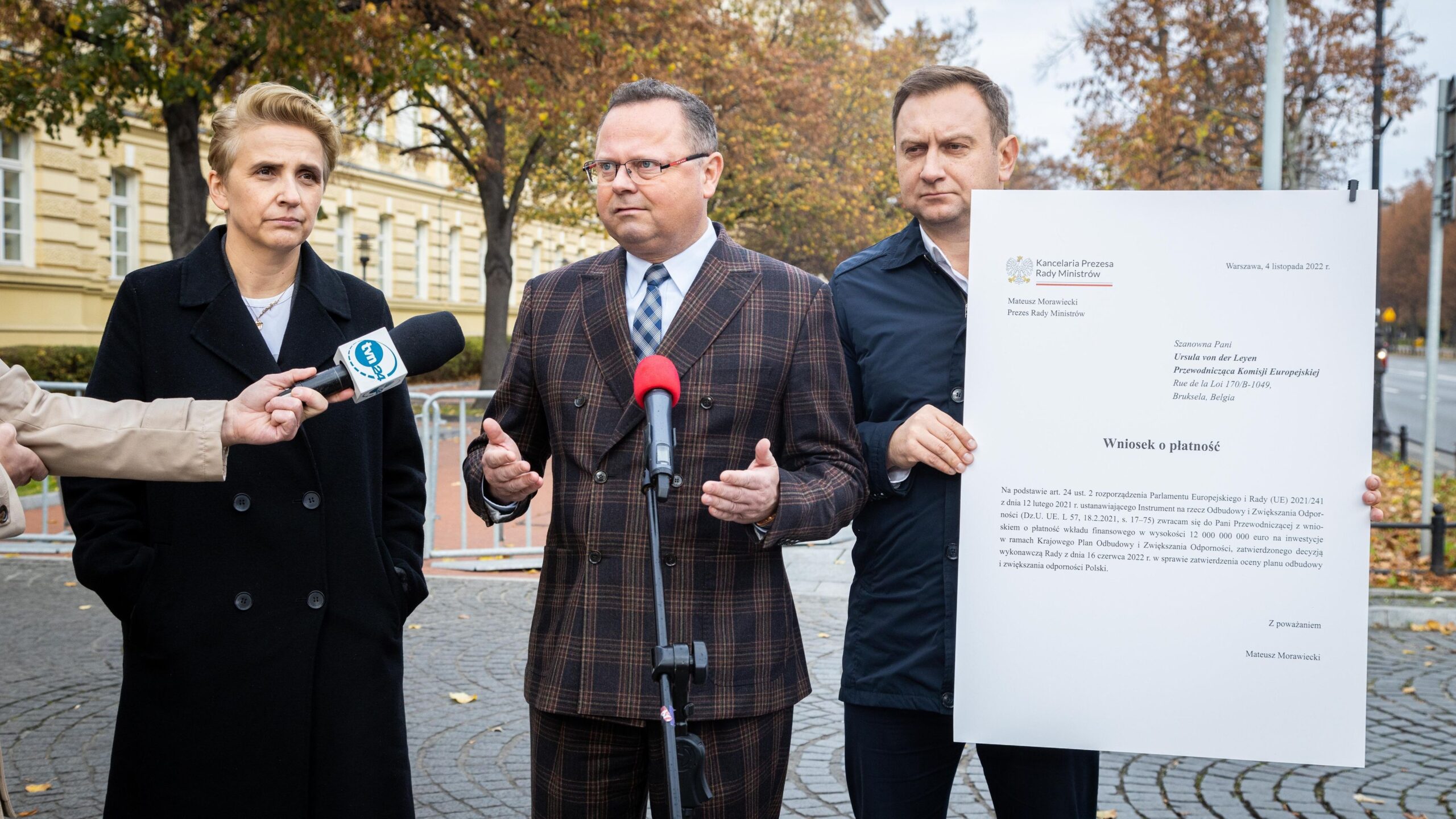 Na zdjęciu widoczna jest konferencja prasowa, podczas której mężczyzna w eleganckim garniturze przemawia do mikrofonu, a obok niego stoi dwóch mężczyzn trzymających dokument oraz kobieta, wszyscy w formalnych ubraniach, na tle jesiennego krajobrazu.