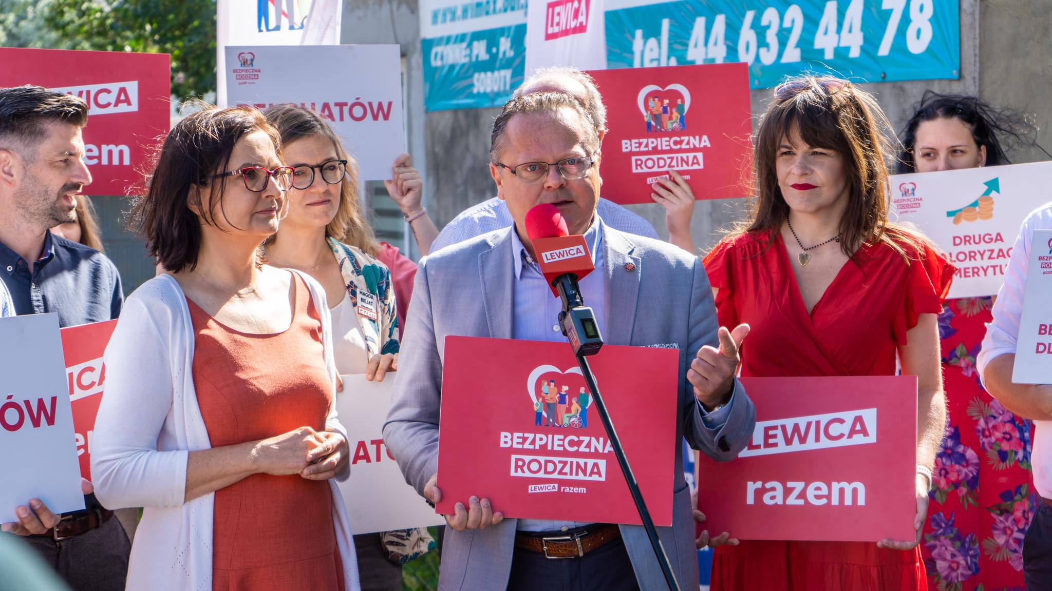 Na zdjęciu widać grupę ludzi, w tym mężczyznę z mikrofonem, prowadzącego konferencję prasową na temat bezpieczeństwa rodzin, przy trzymanych przez uczestników transparentach związanych z kampanią polityczną.