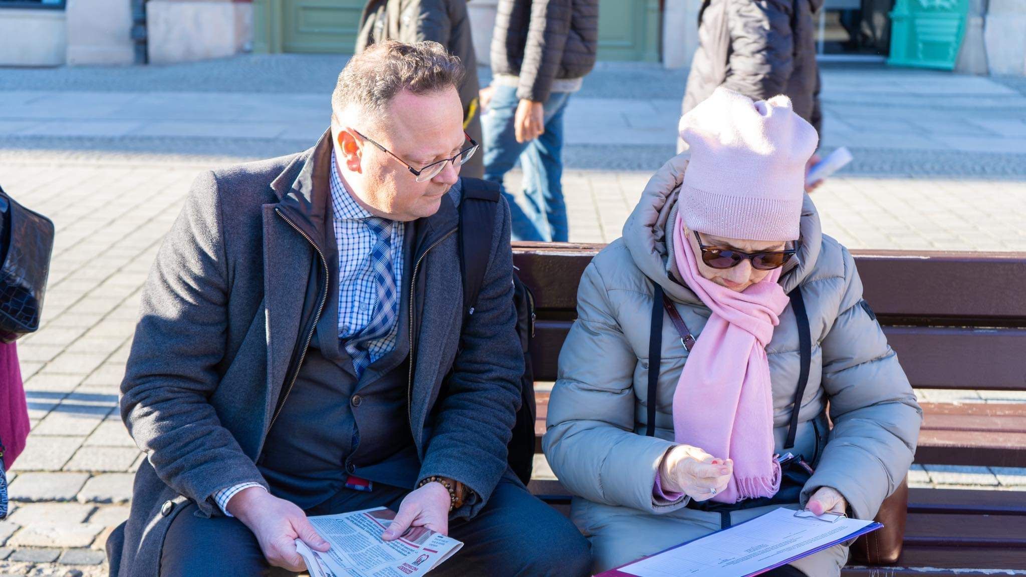 Dwóch dorosłych siedzi na ławce na zewnątrz: mężczyzna w granatowym płaszczu przegląda gazetę, a kobieta w jasnoszarej kurtce, różowym szaliku i czapce czyta dokumenty.