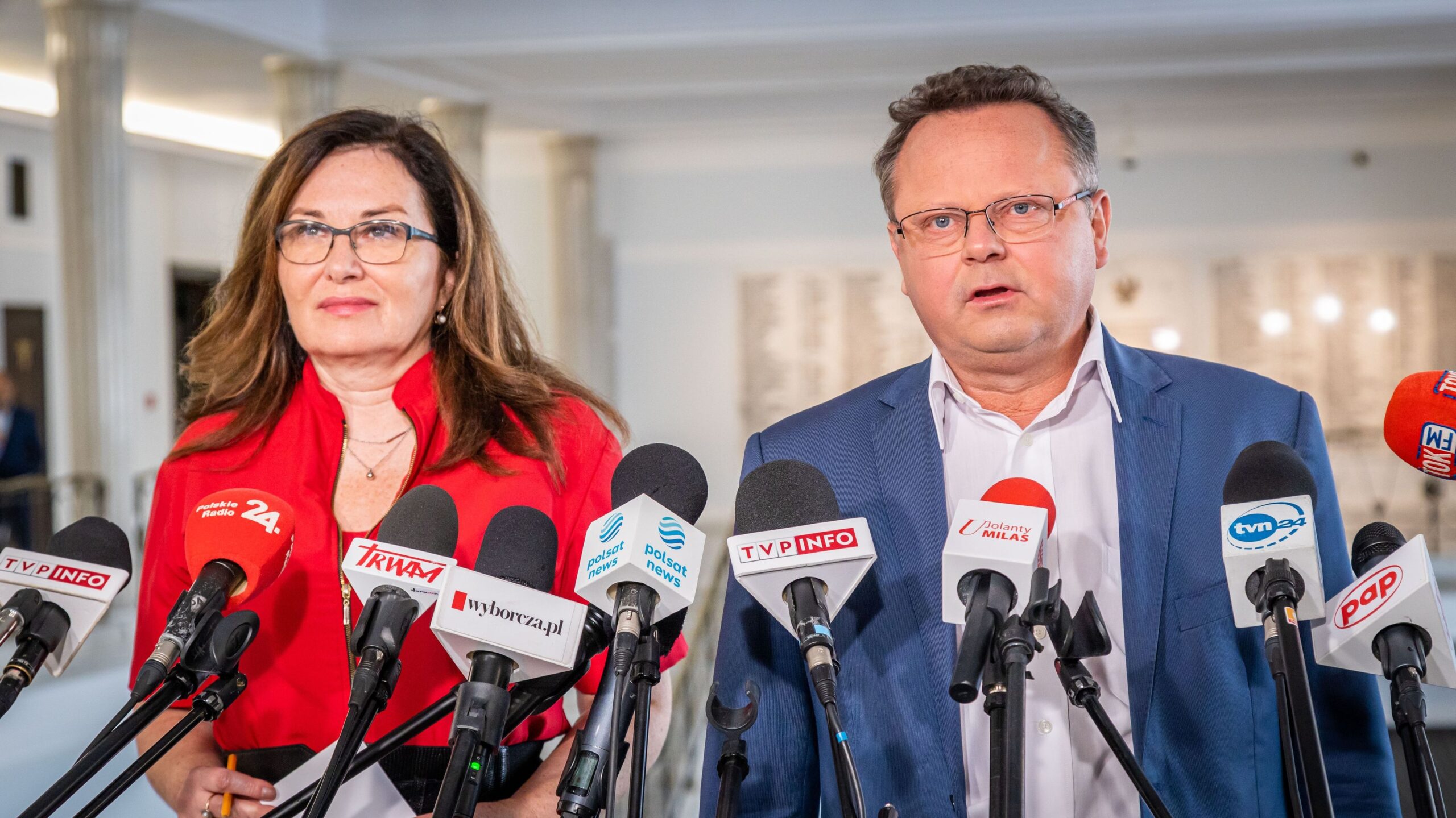 Dwie osoby stoją przy licznych mikrofonach podczas konferencji prasowej.