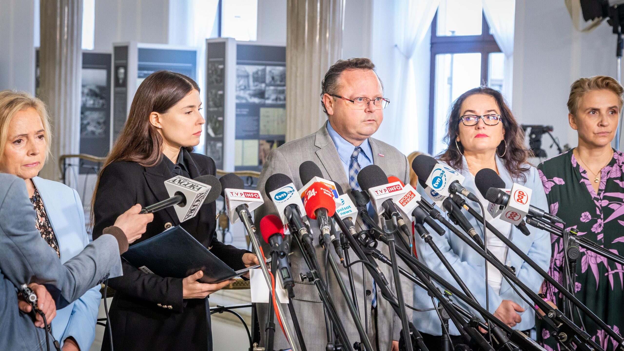 Na zdjęciu grupa osób stoi przed mikrofonami podczas konferencji prasowej w jasnym pomieszczeniu.