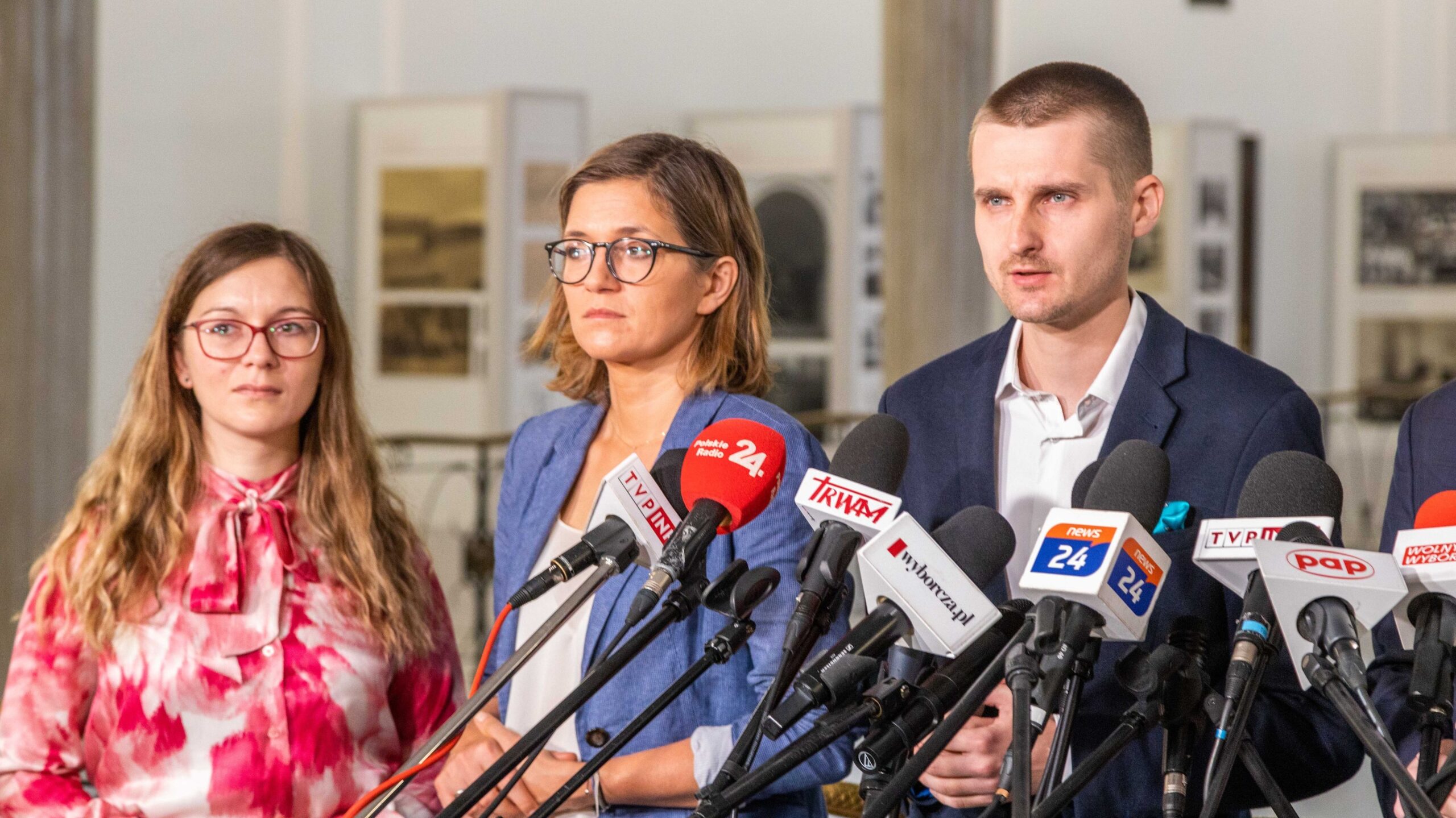 Na zdjęciu widoczna jest trójka ludzi podczas konferencji prasowej, stojących przed mikrofonami i aparatami medialnymi.